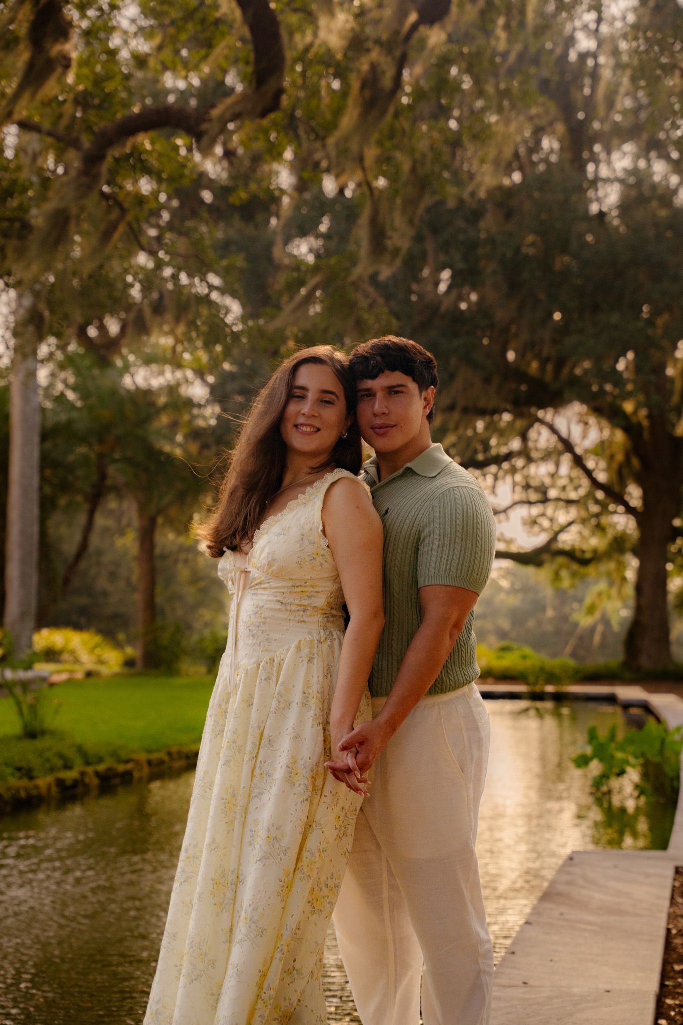 Elegant couple holding hands on a wooden dock surrounded by trees and a reflective pond at sunset.