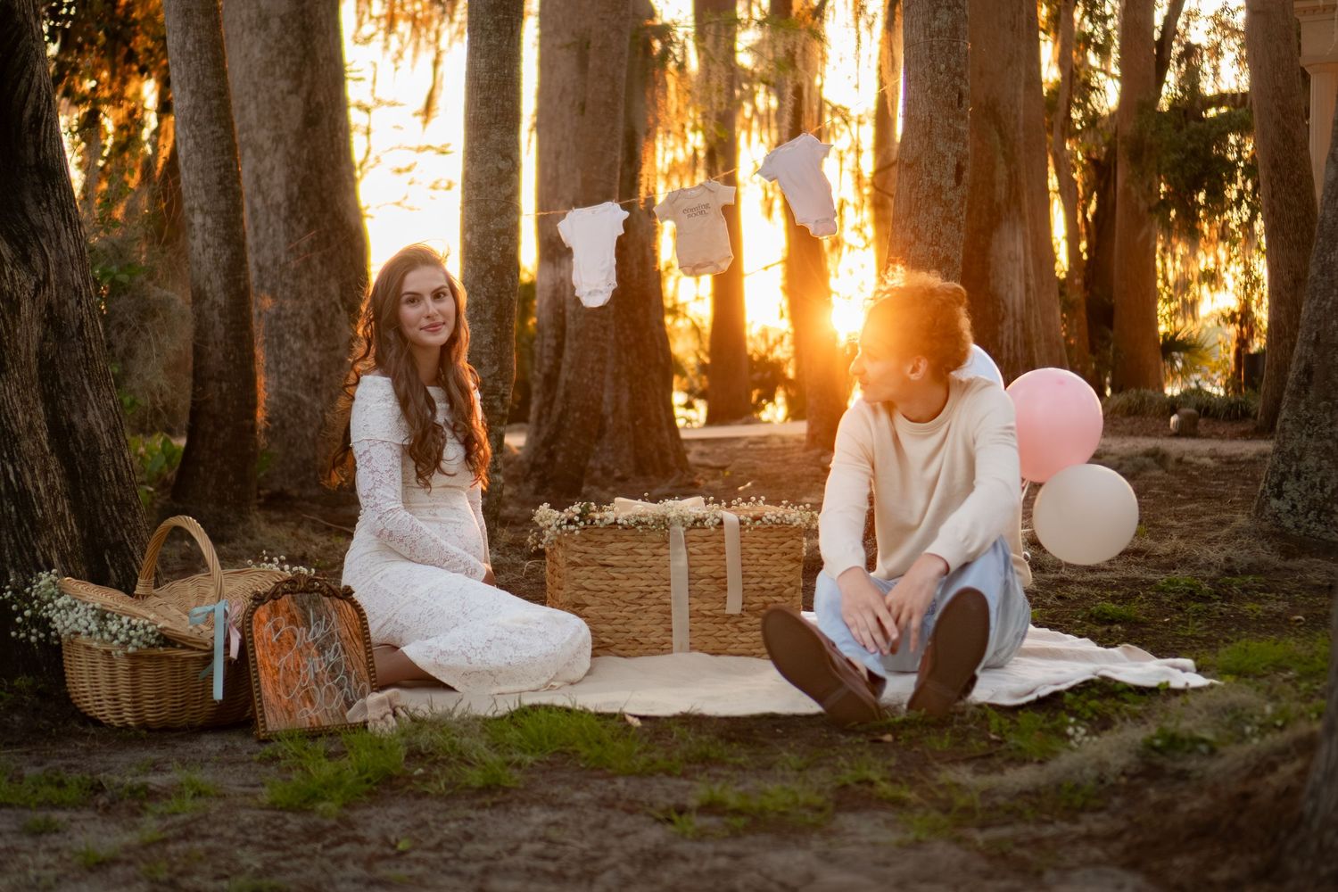 Expecting couple enjoying a woodland picnic at sunset with balloons, basket, and baby decorations.