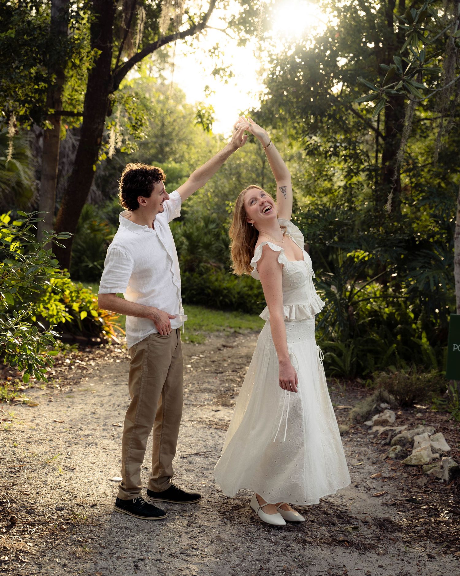 A joyful couple dancing on a sunlit forest path, the woman in a white dress being twirled by her partner.