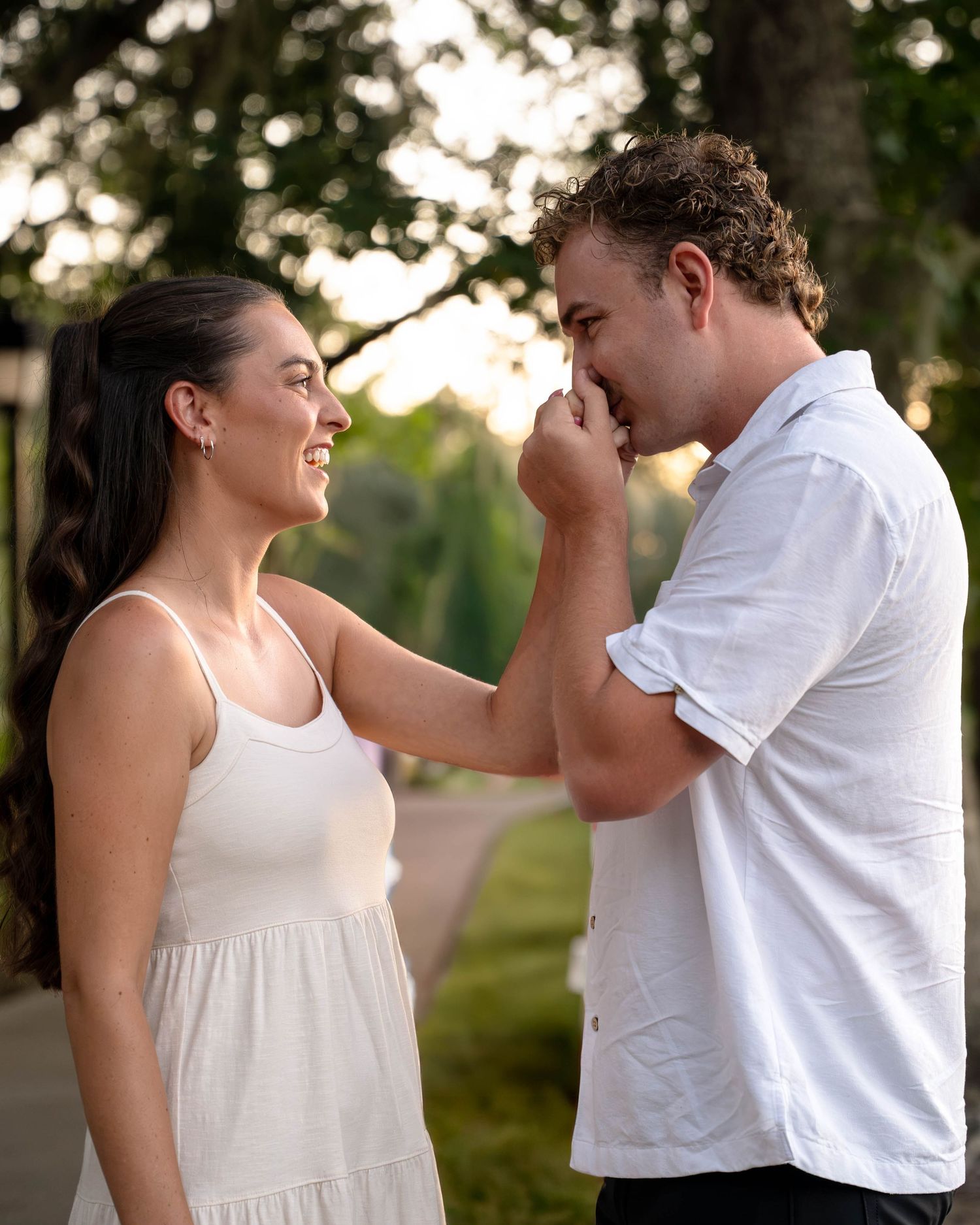 A romantic couple in white outfits share a tender moment outdoors, the man gently kissing the woman's hand.