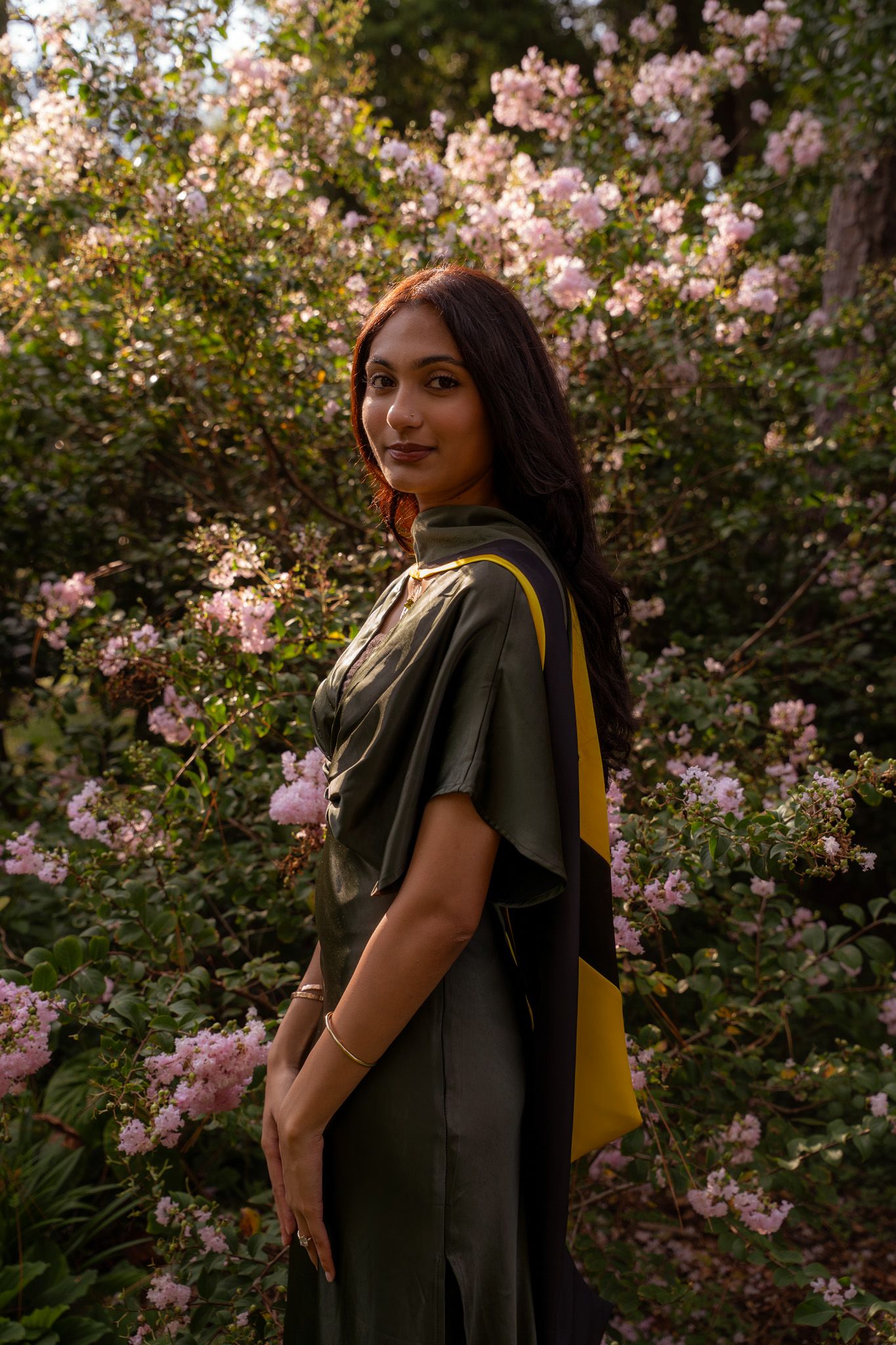 Young woman in dark graduation gown with yellow sash stands among blooming pink flowers in garden.