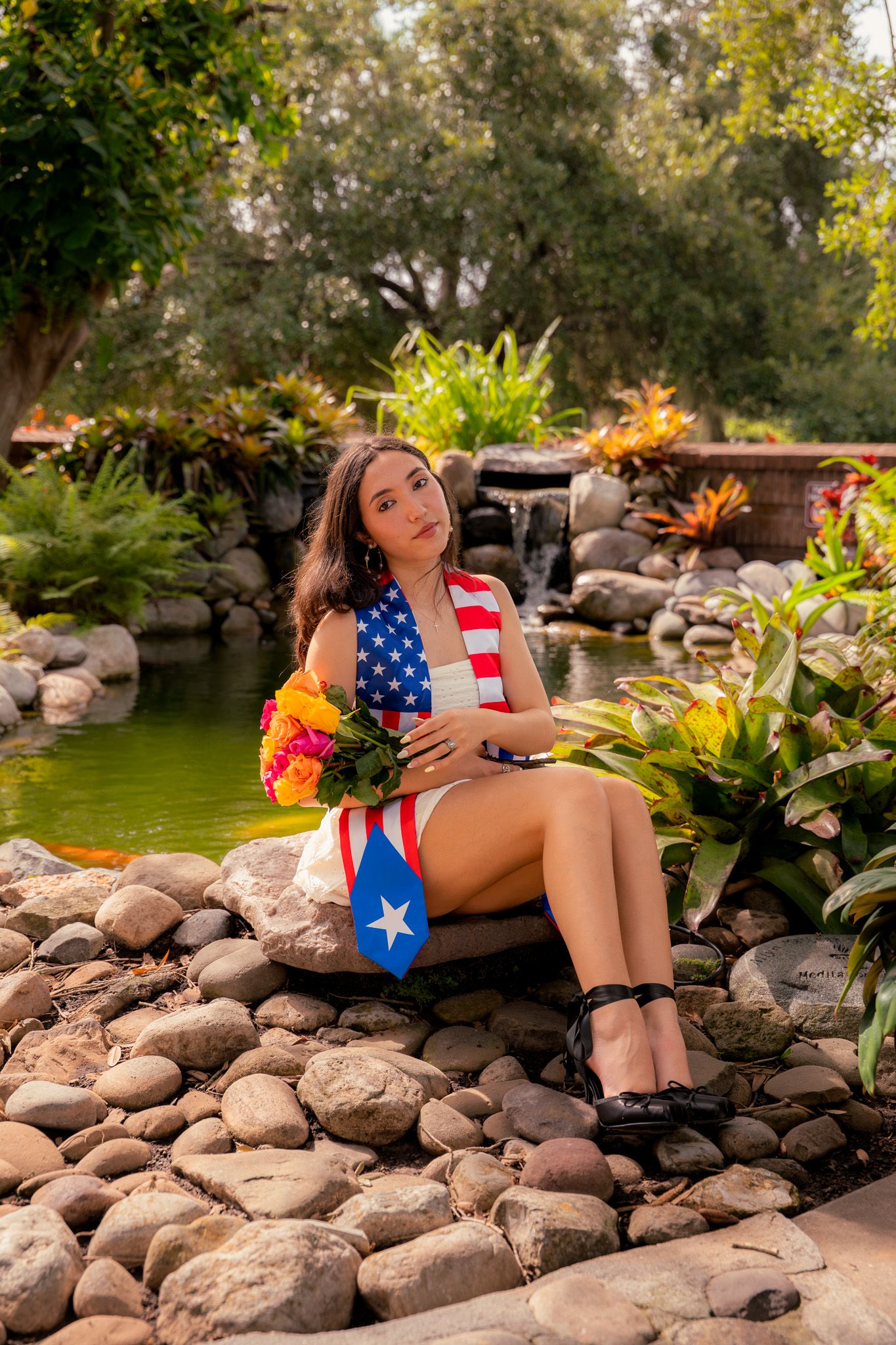 Young woman in white dress draped with American and Puerto Rican flags sits by garden pond.
