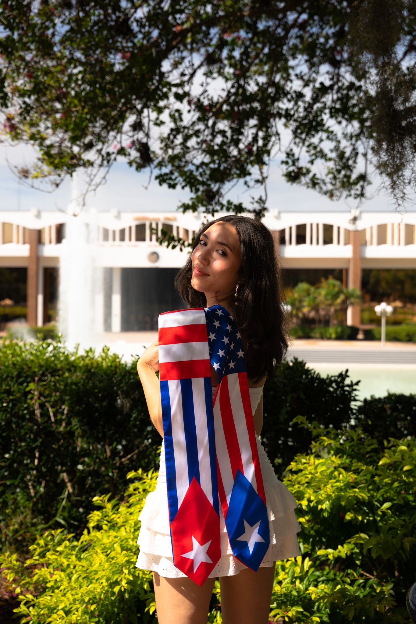 Young woman wrapped in American and Puerto Rican flags stands in lush campus garden.