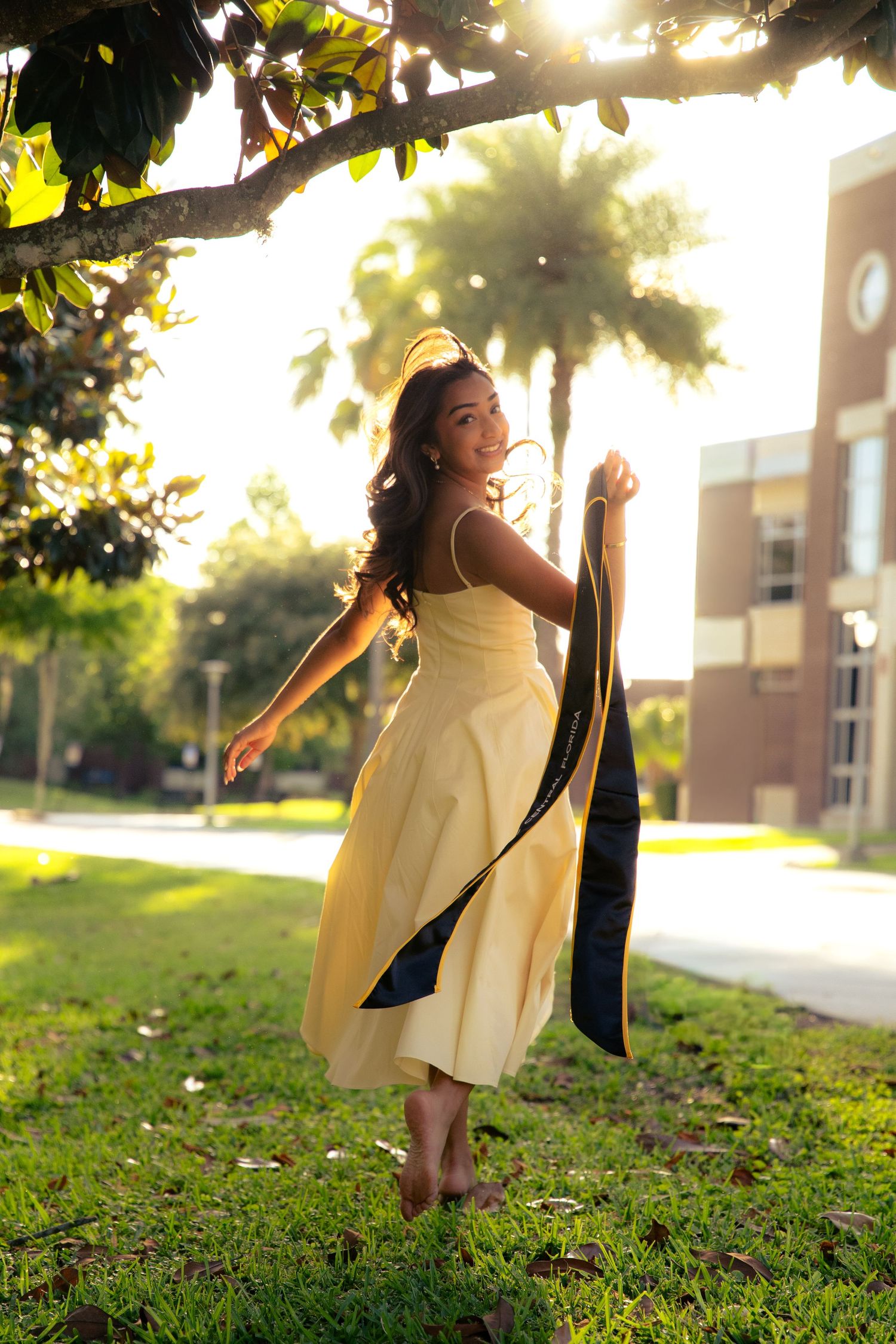 Woman in yellow dress twirls holding graduation gown under tree in golden sunset light.