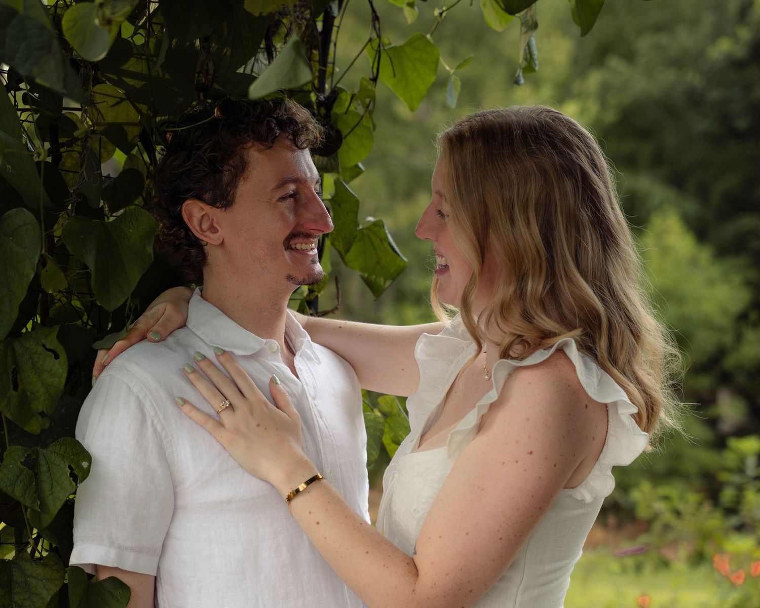 A smiling couple stands close together under leafy branches in an outdoor engagement photo.