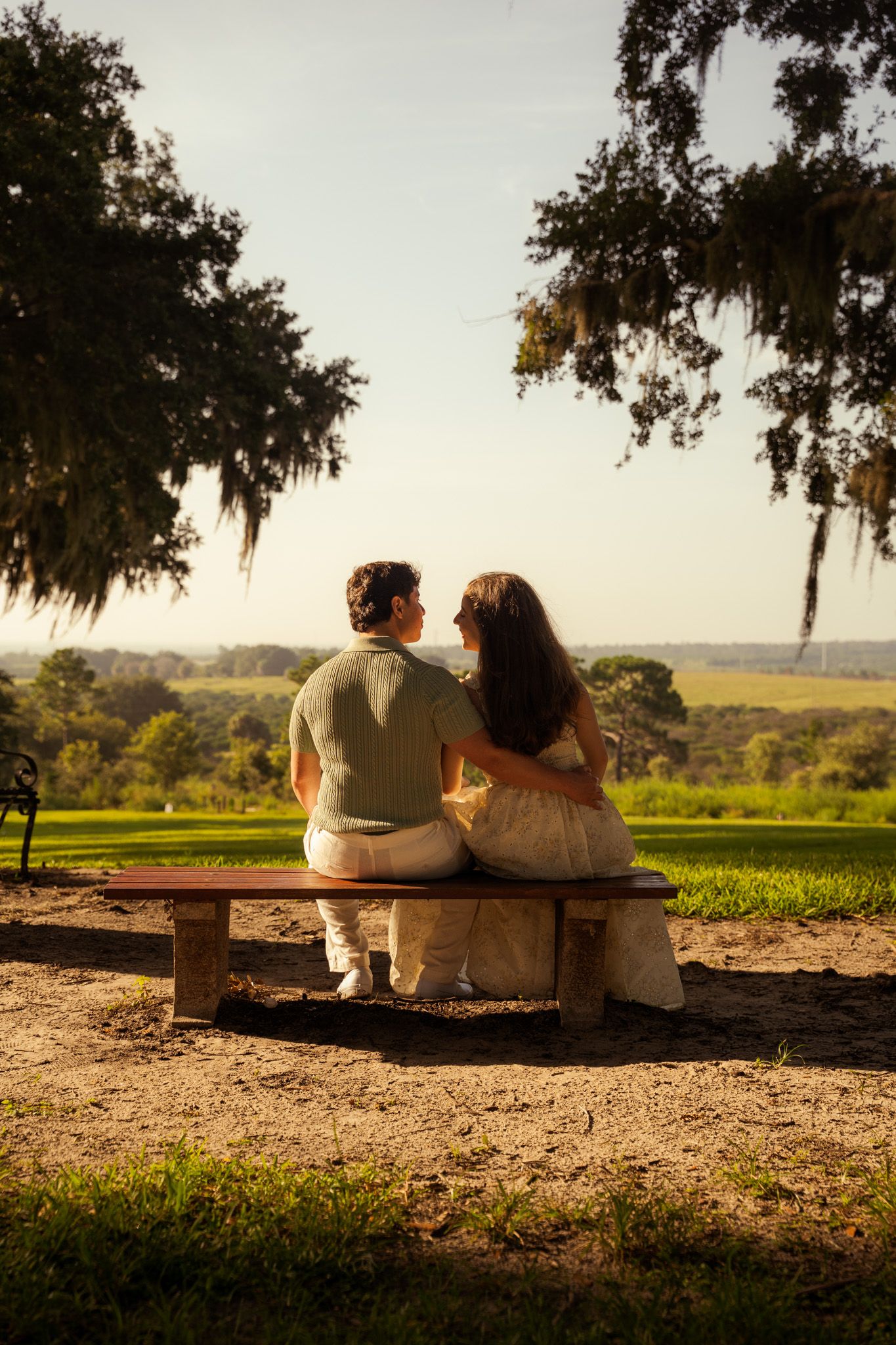 Romantic couple sitting together on a wooden bench overlooking a lush green landscape at golden hour.