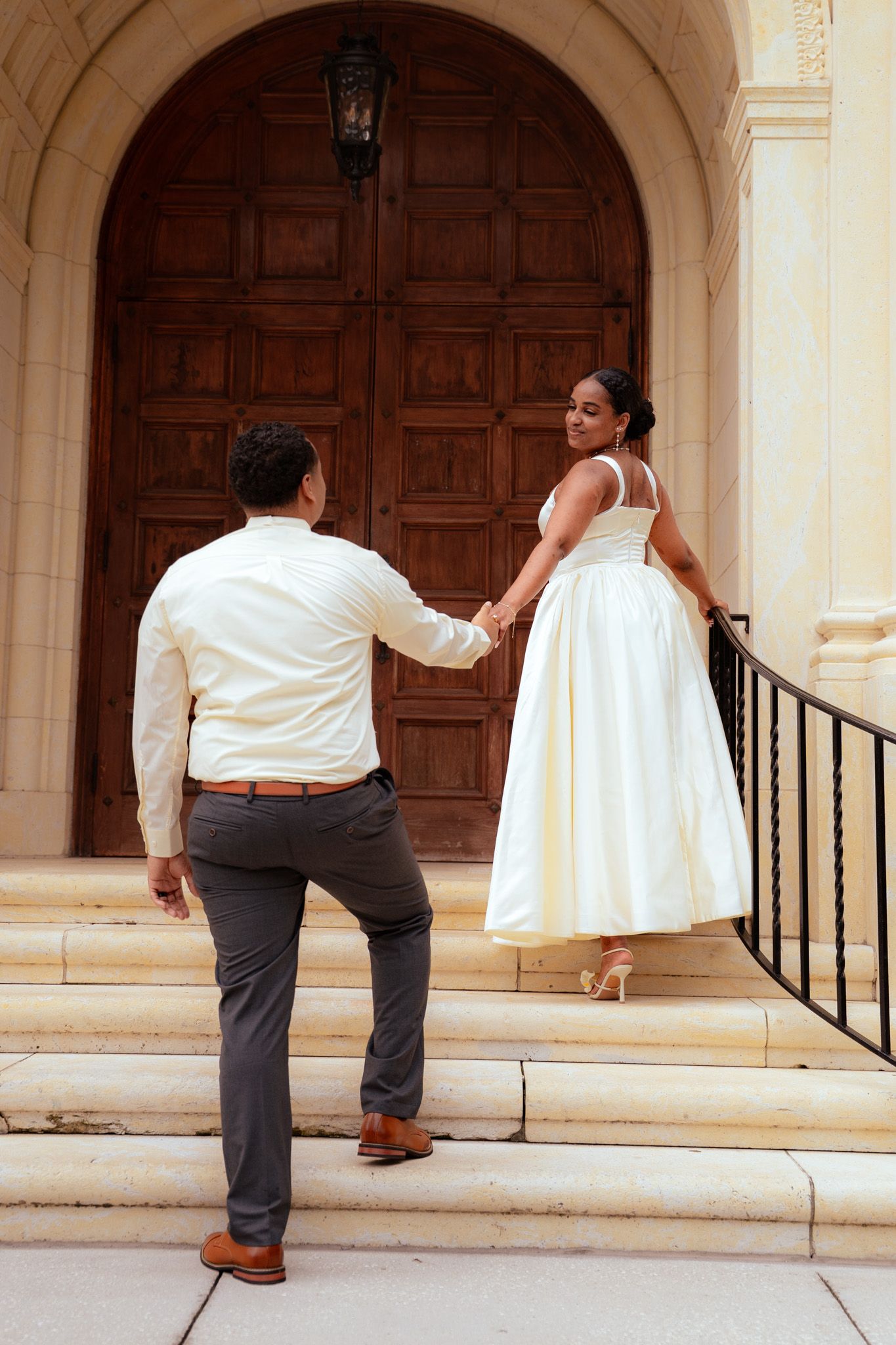 Bride in white midi dress leads groom by hand up steps to ornate wooden church doors.