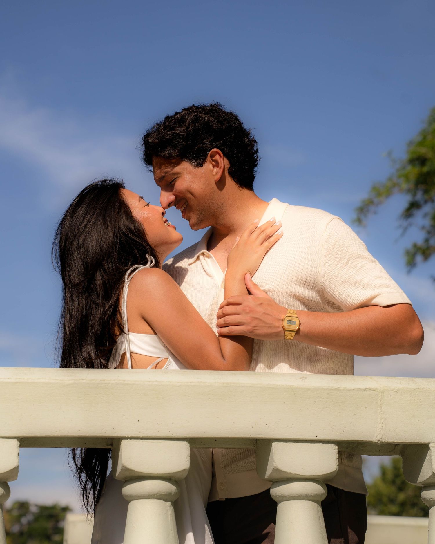A joyful couple leans in close together on a white balcony under a blue sky.