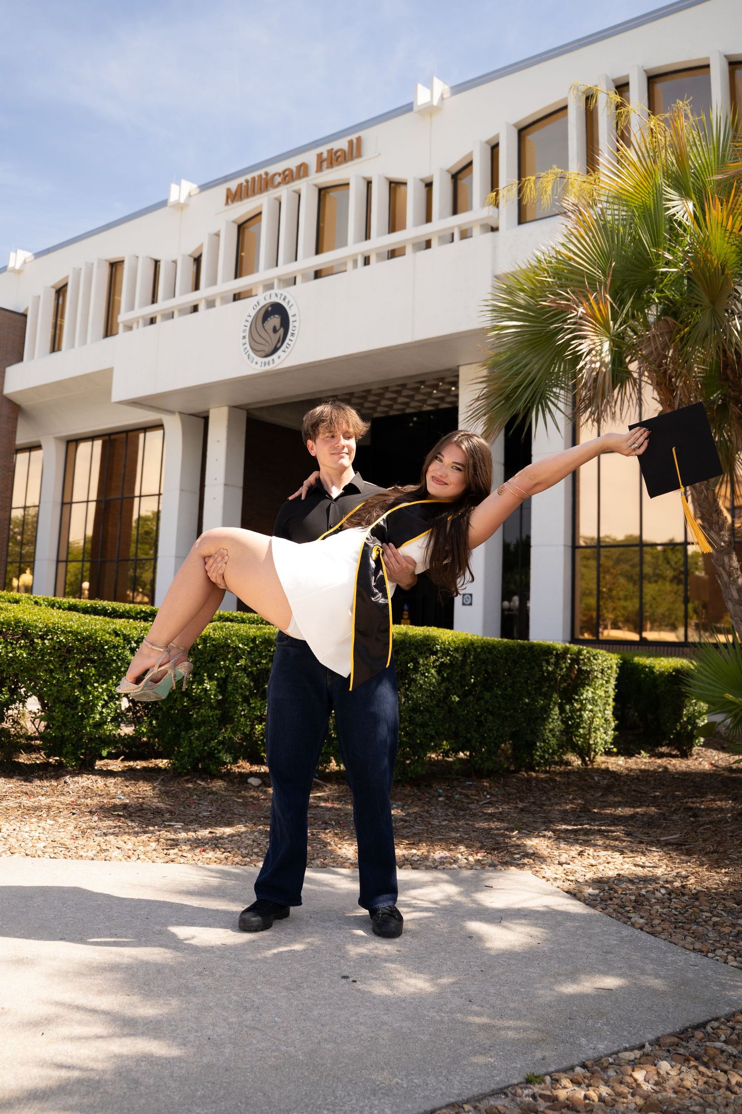 Graduate in cap and gown being lifted by partner in front of Millican Hall building on sunny day.