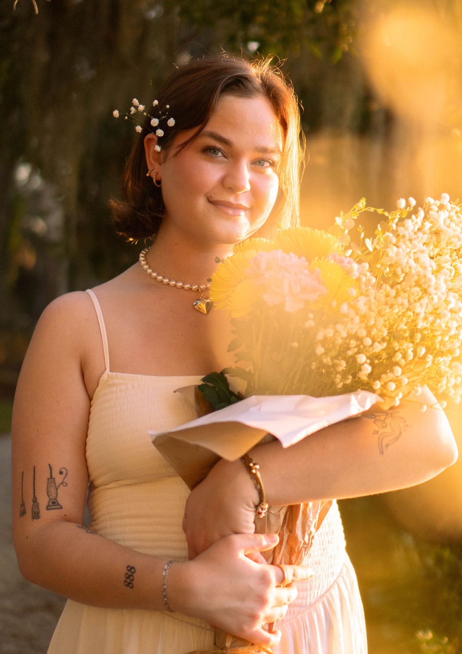 Young woman in cream dress holding yellow bouquet with baby's breath hair accessory and pearl necklace in golden sunlight.