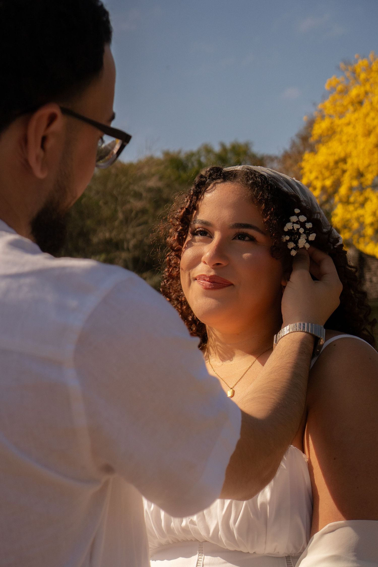 Man tenderly adjusting bride's floral hair accessory outdoors with yellow blooms in sunny background.
