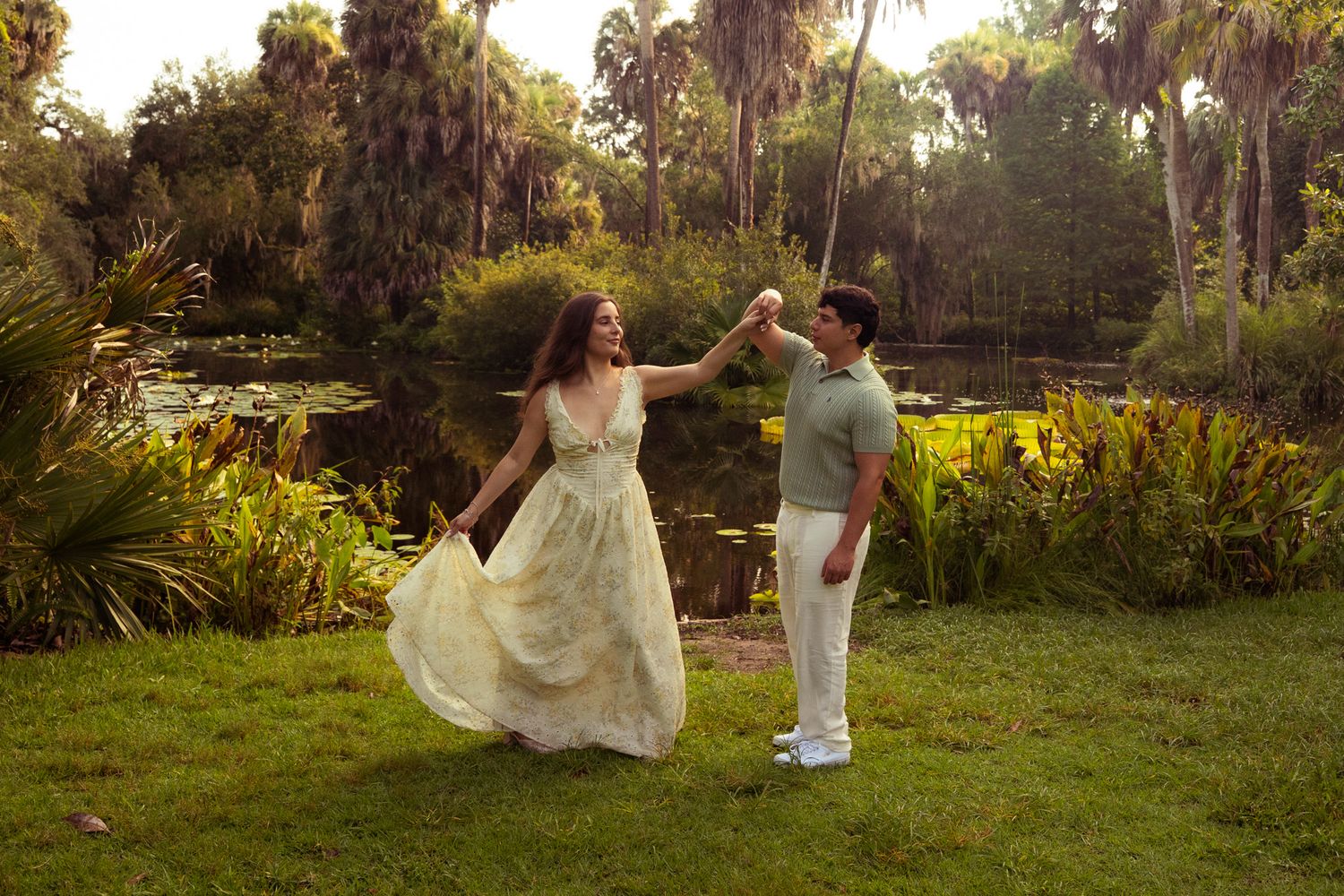 Joyful couple dancing together on green grass beside a scenic pond surrounded by tropical plants.