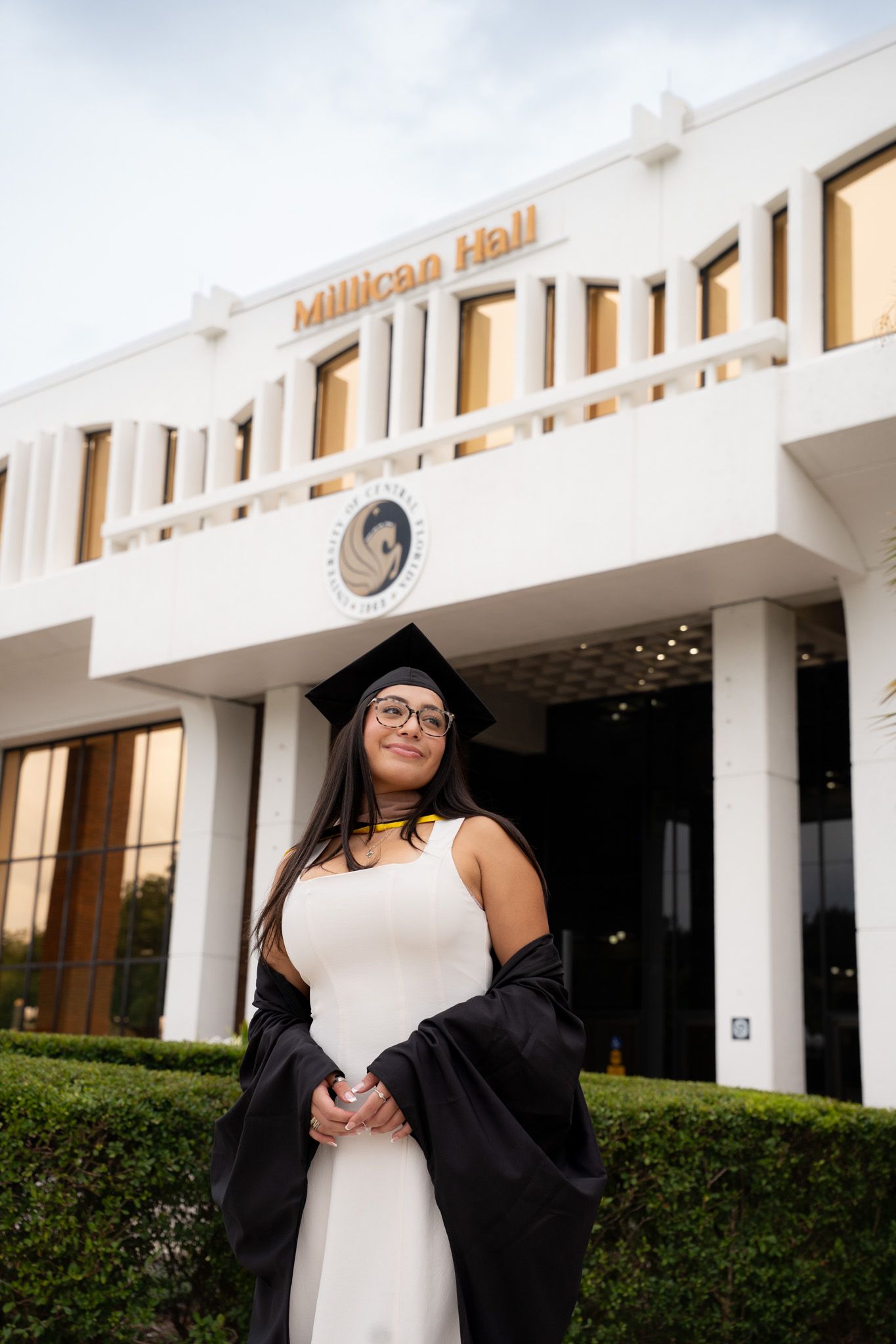 Female graduate in cap and gown stands proudly before Millican Hall building in white dress and glasses.