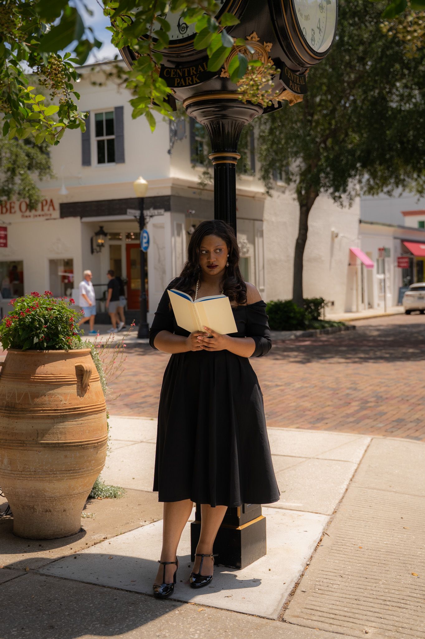 Woman in black off-shoulder dress holding a book while standing by a decorative street lamp.