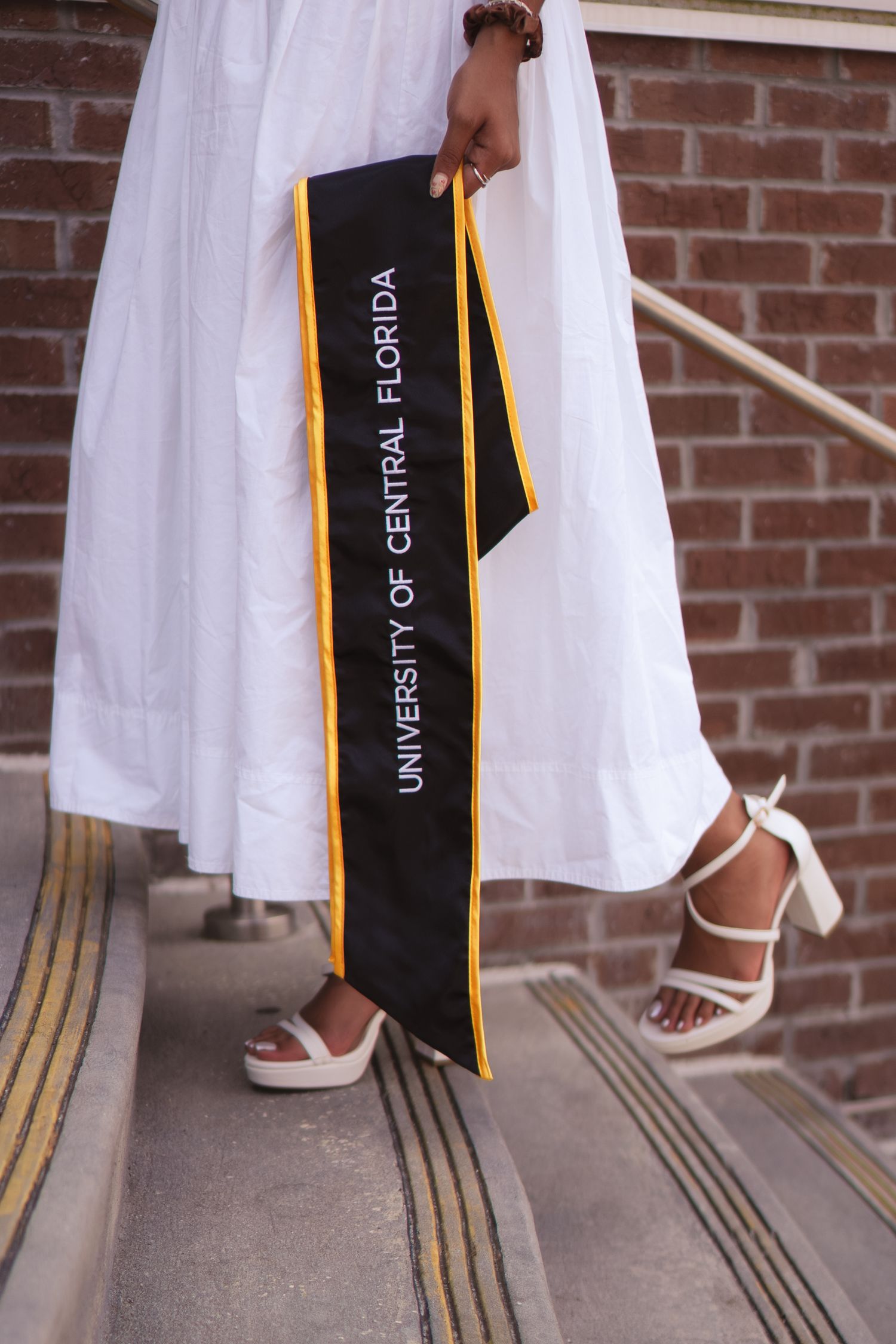Close-up of woman in white dress holding University of Central Florida graduation sash on steps.