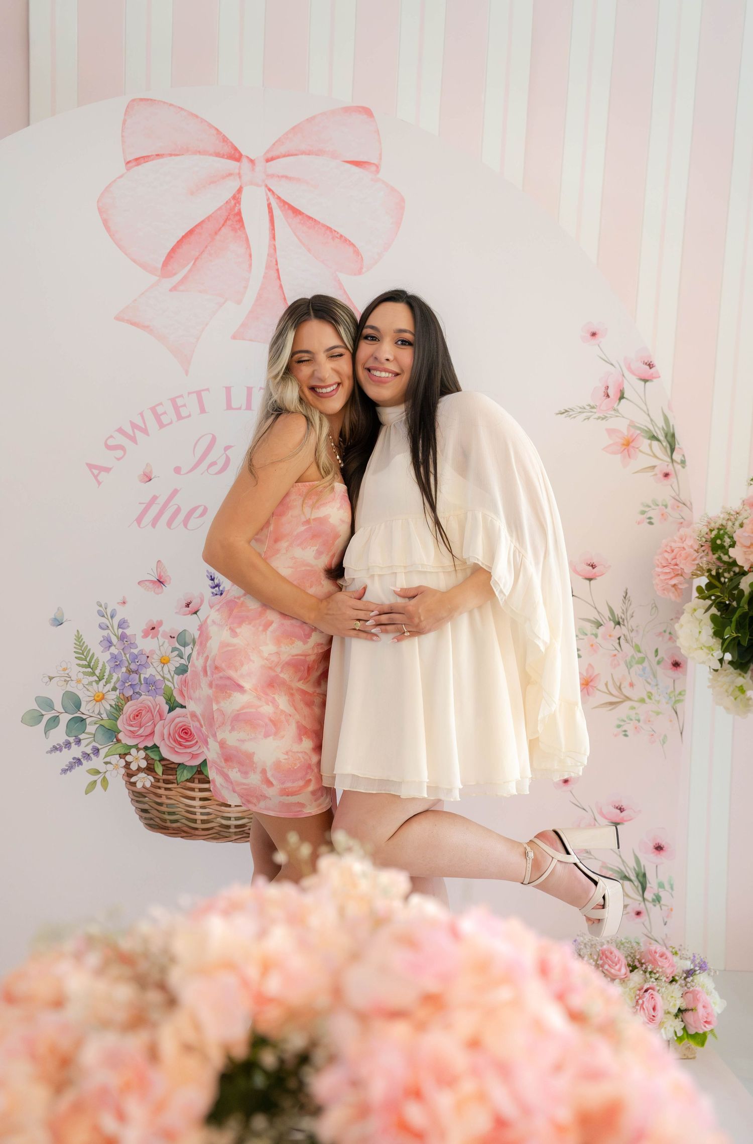 Two women posing together at a pink baby shower backdrop, one pregnant in white dress.
