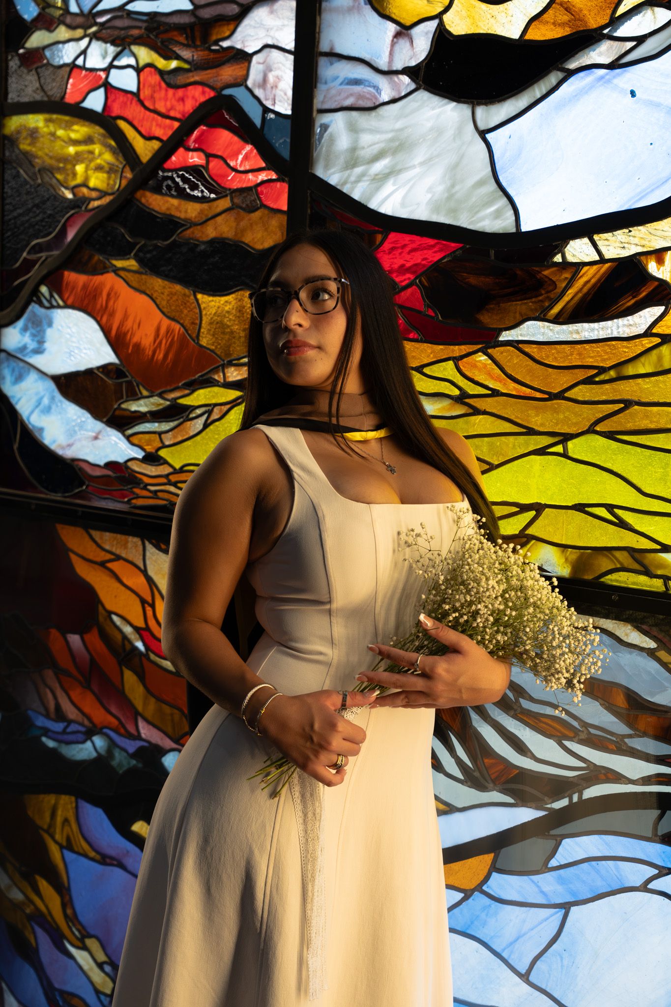 Young woman in white dress holding baby's breath flowers before a colorful stained glass window backdrop.