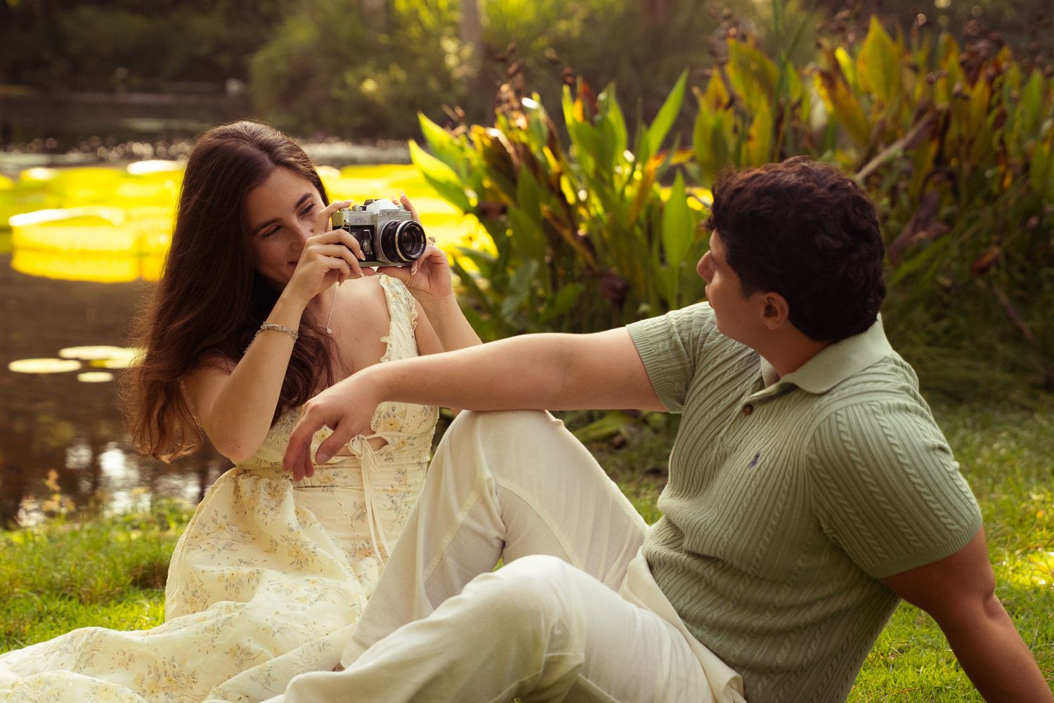 Couple sitting on grass near a pond, woman holding a vintage camera while man reaches toward her.