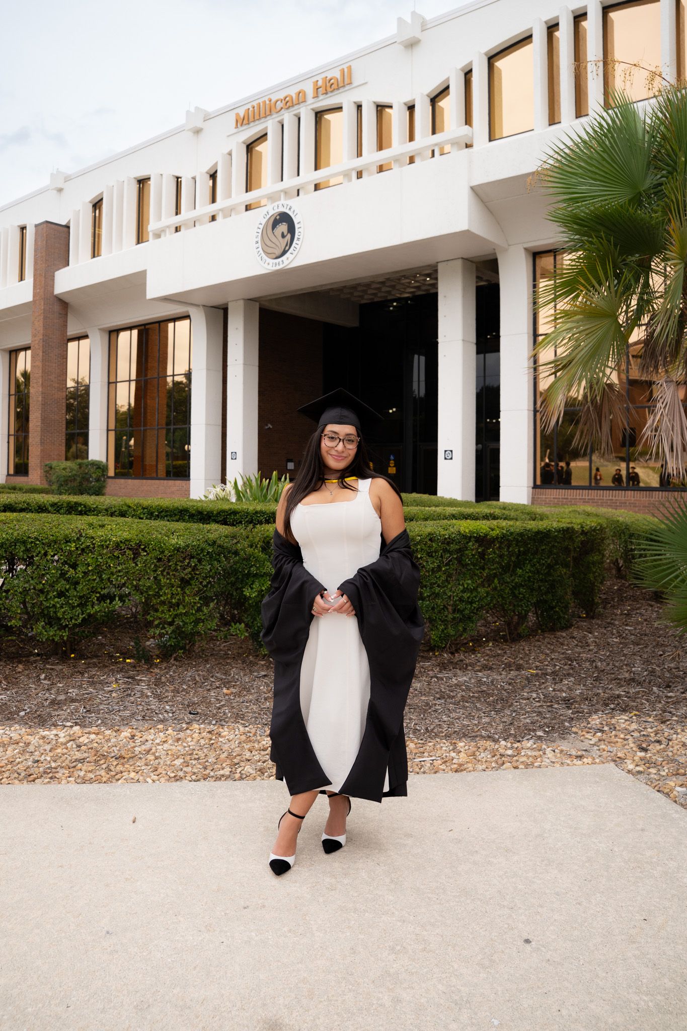 Female graduate in black cap and gown poses in white dress before Millican Hall university building.
