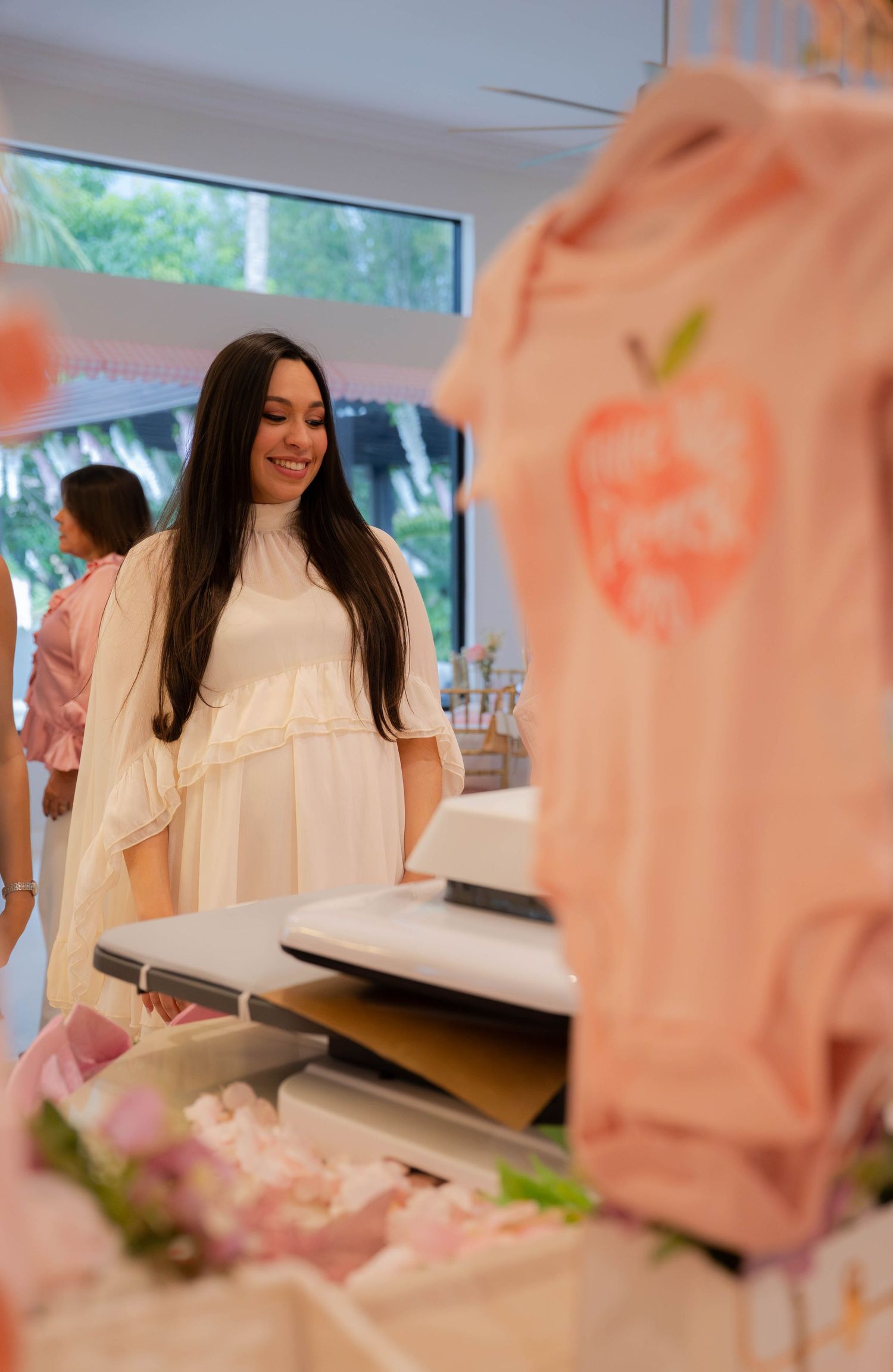 Pregnant woman in white dress smiling at pink baby girl clothing display at baby shower.