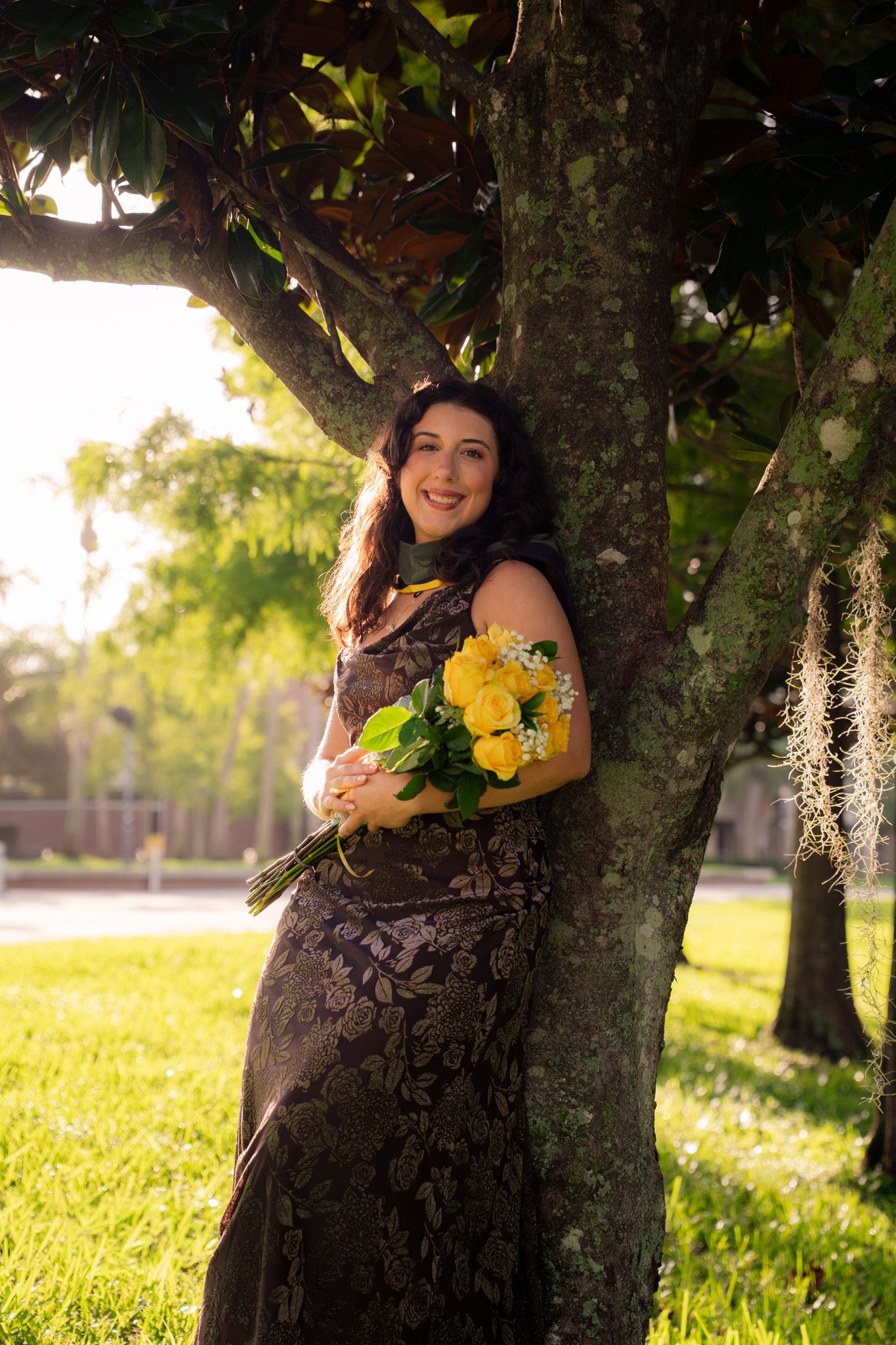 Smiling woman in dark floral gown holds yellow rose bouquet leaning against sunlit tree.