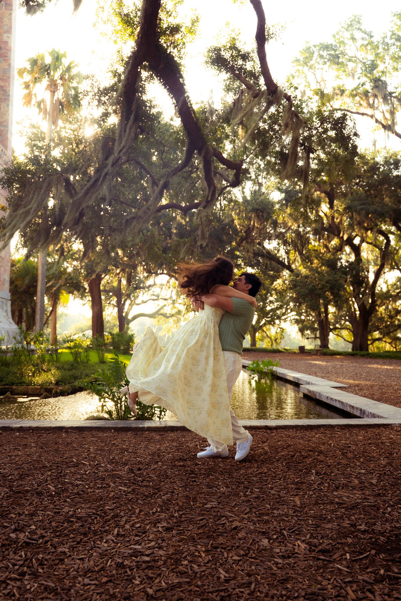 Man lifts woman in yellow floral dress near reflective pond surrounded by sunlit oak trees.