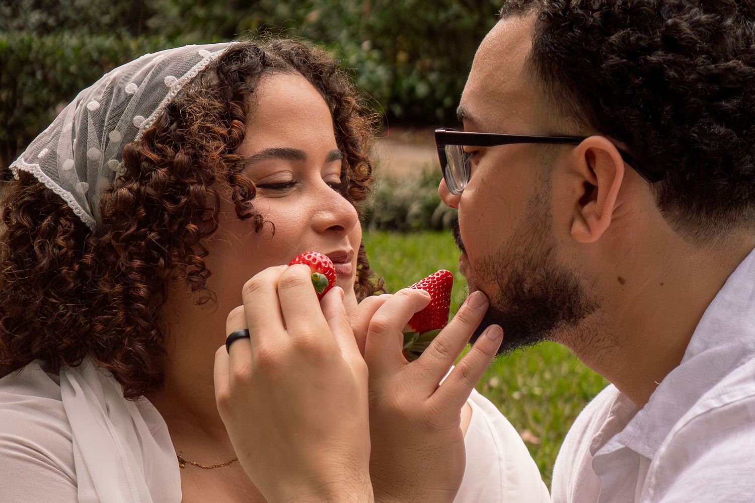 Couple sharing strawberries outdoors in a romantic moment, woman wearing ring with curly hair, man in glasses.