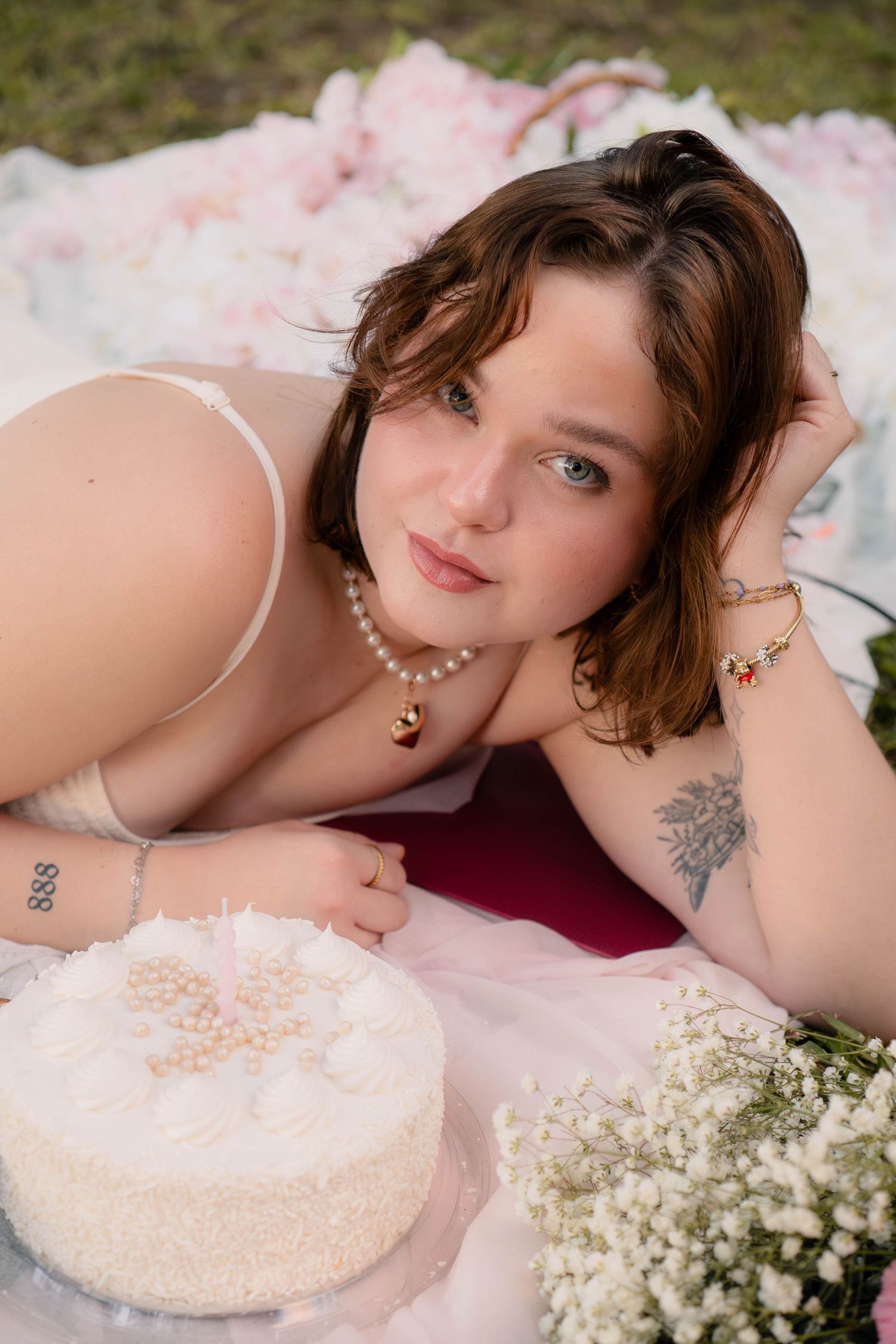 Young woman in lingerie lying on a blanket with a white cake and flowers in an outdoor setting.
