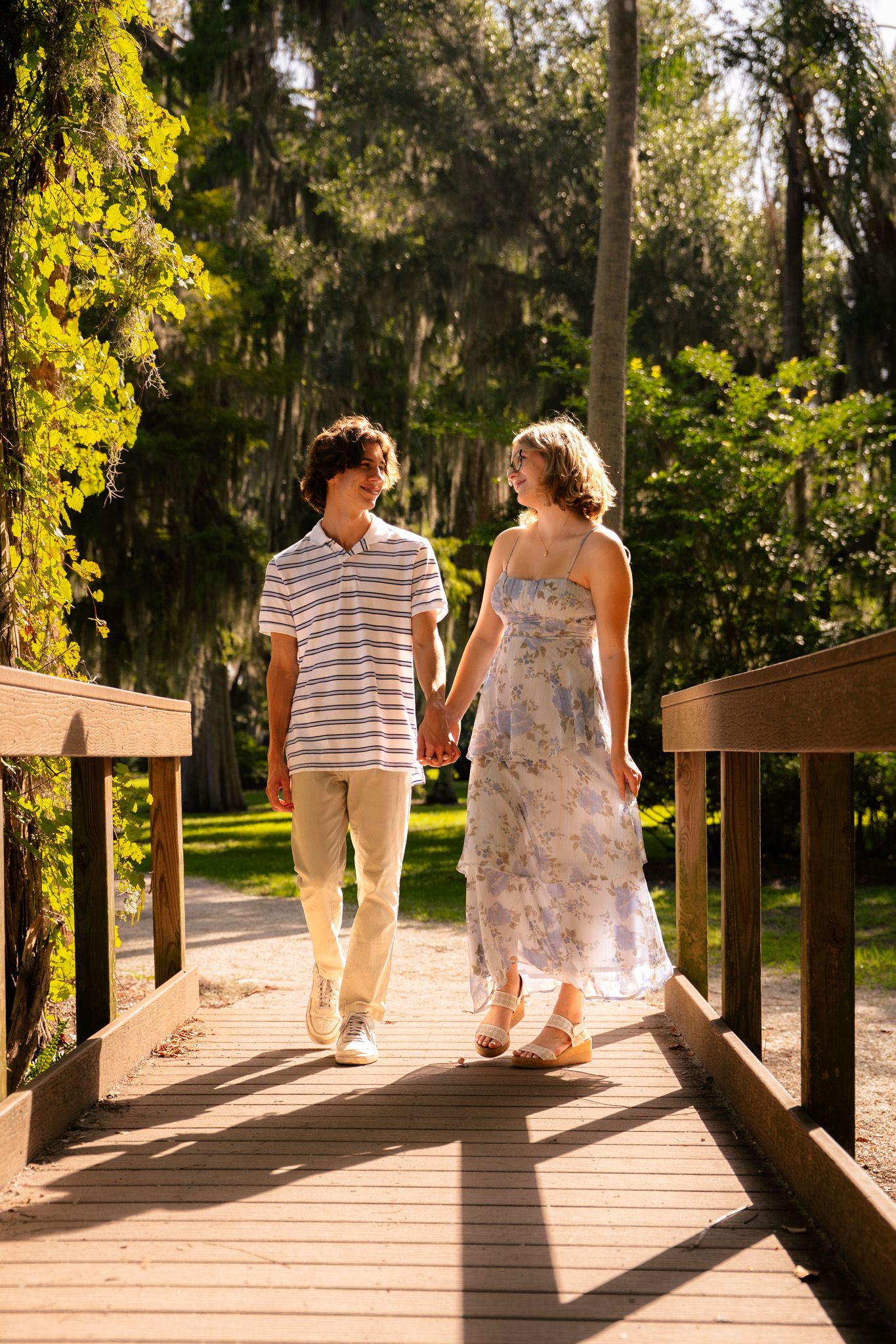 A young couple holds hands while walking on a sunny wooden bridge surrounded by lush green trees.