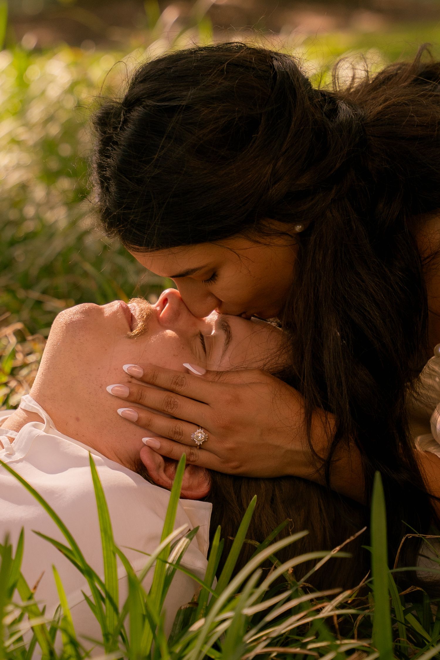 Woman with engagement ring kisses man lying in grass during golden hour outdoor photo session.
