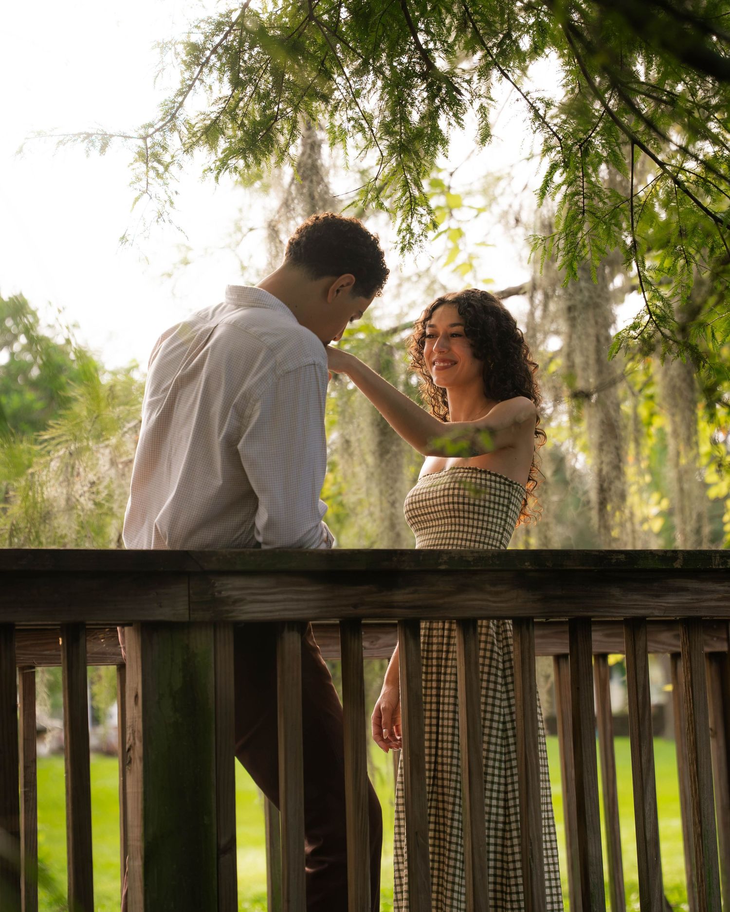 Couple sharing a romantic moment on a wooden deck surrounded by lush green trees in soft natural light.
