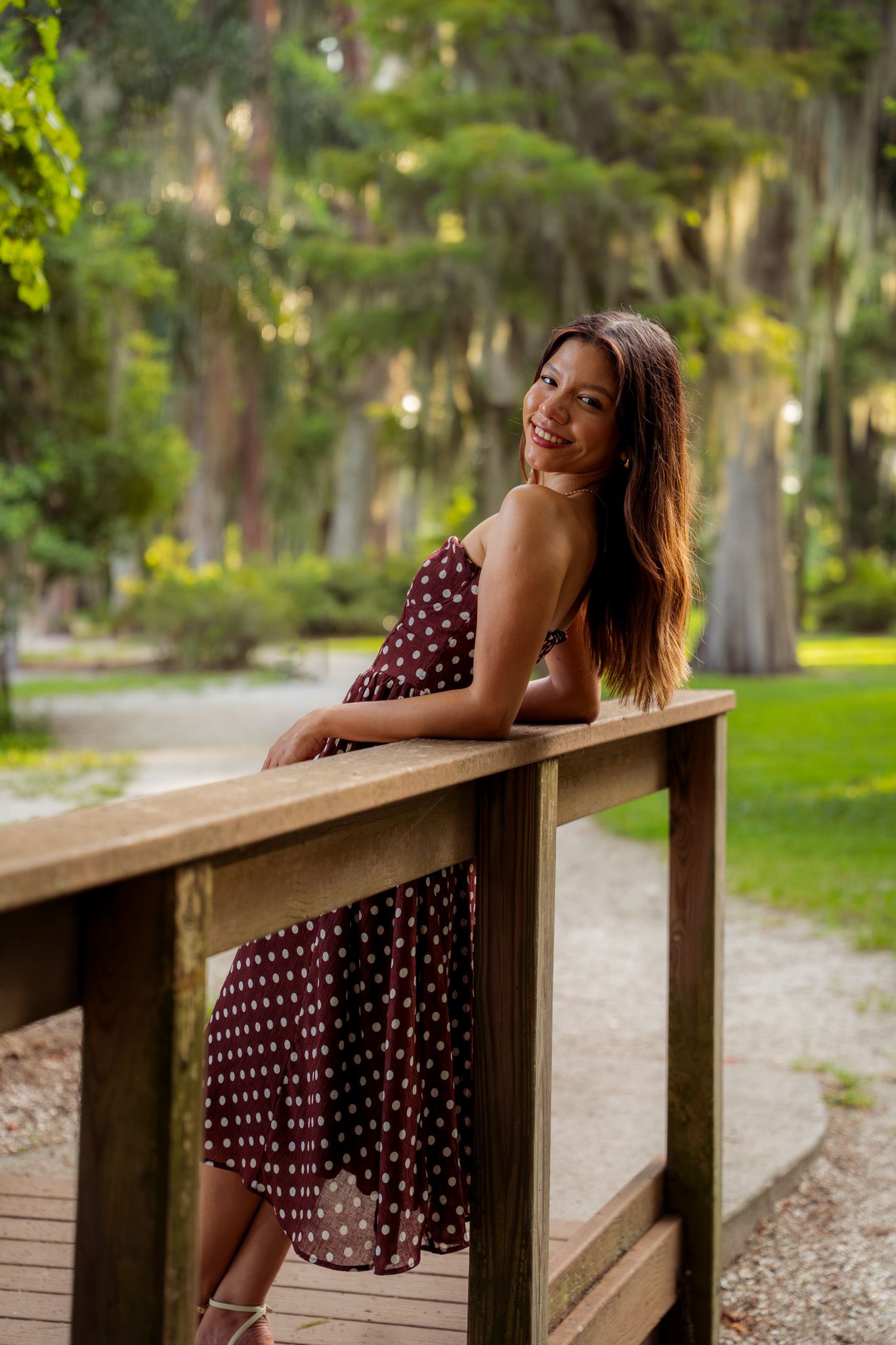Smiling woman in polka dot dress leaning on wooden bridge railing in scenic park with Spanish moss trees.