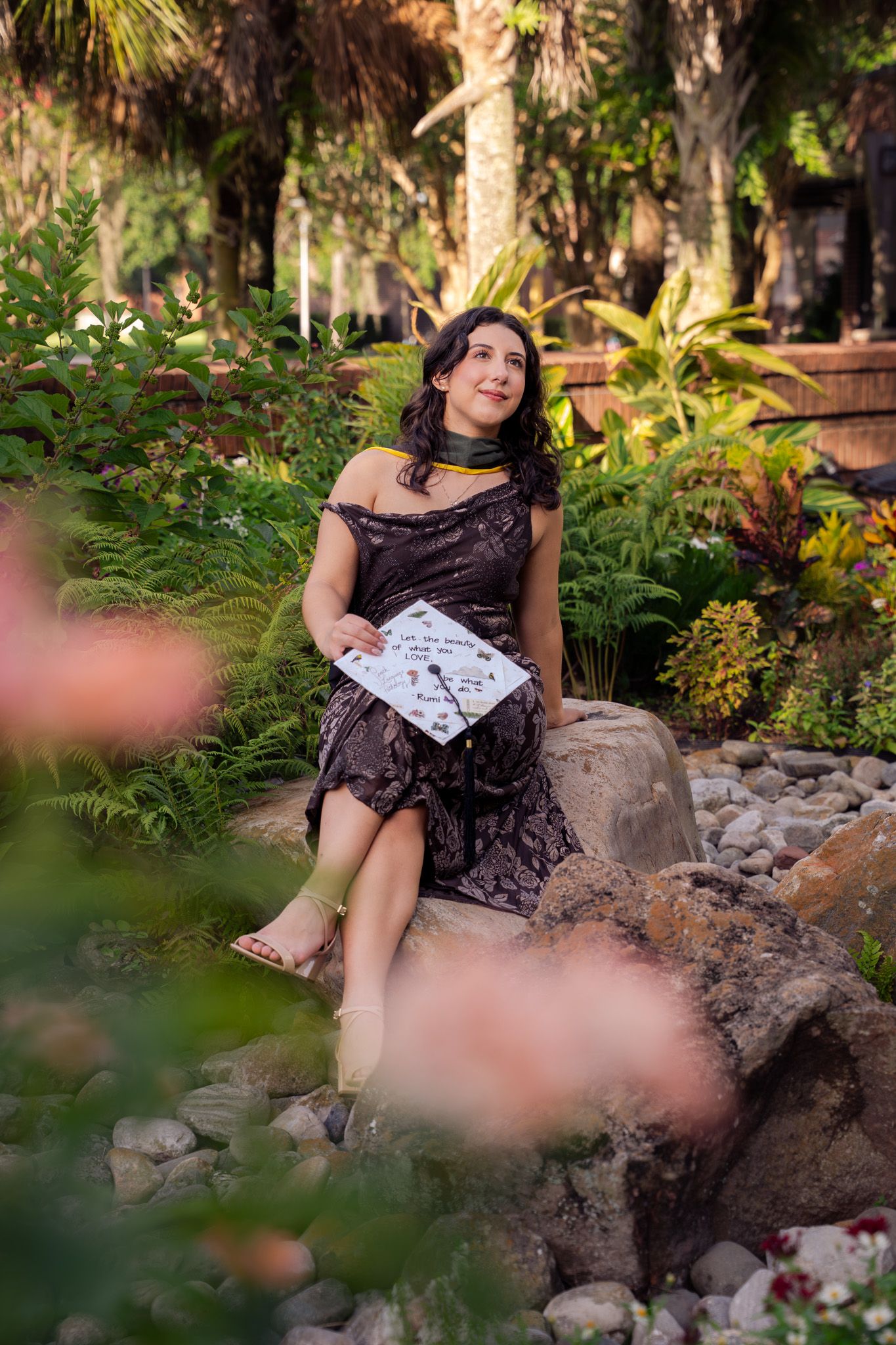 Young woman in brown dress sits on rocks holding decorated graduation cap in lush tropical garden.