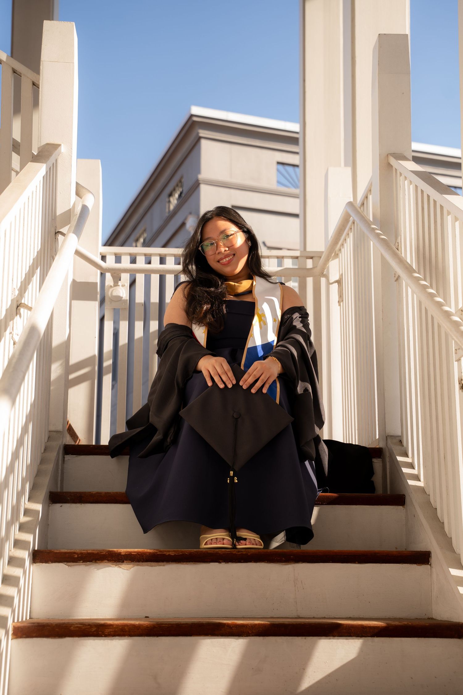 Smiling graduate in black cap and gown sits on white outdoor staircase under blue sky.
