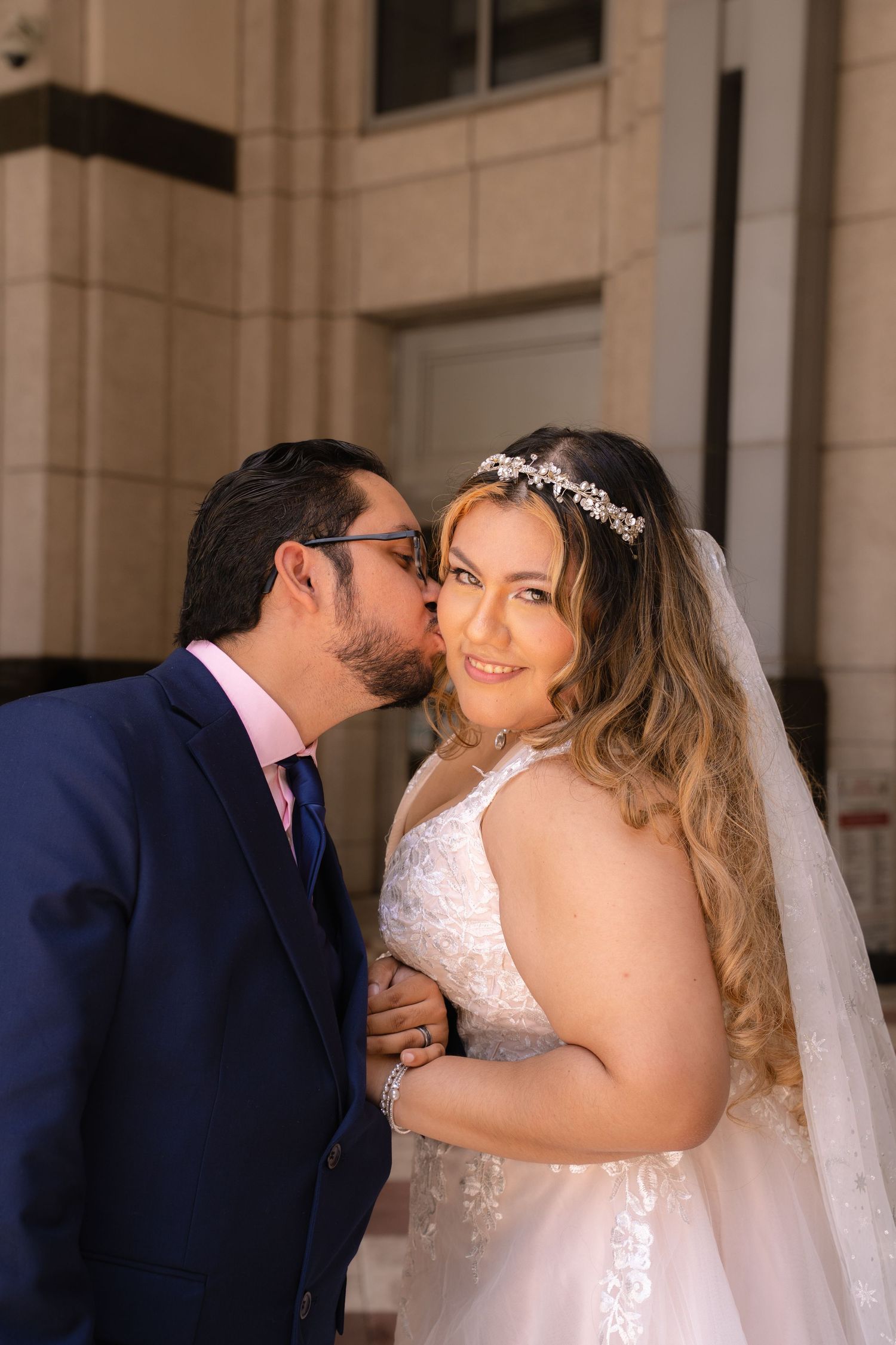 Groom in navy suit kisses bride wearing lace wedding dress and crystal headpiece on cheek.