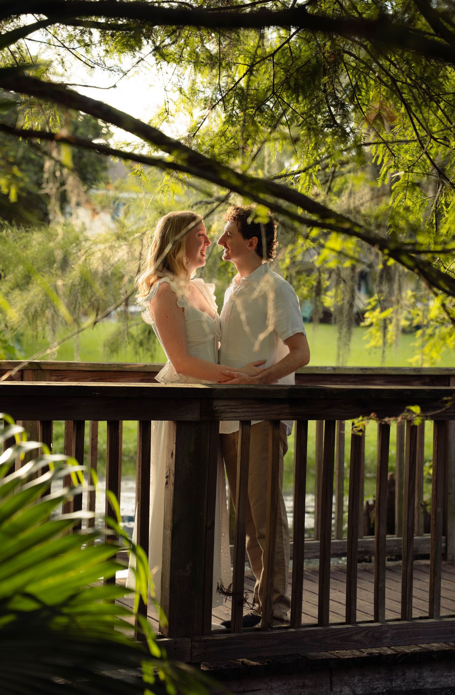 Couple embracing on a wooden deck surrounded by sunlit green trees by a pond.