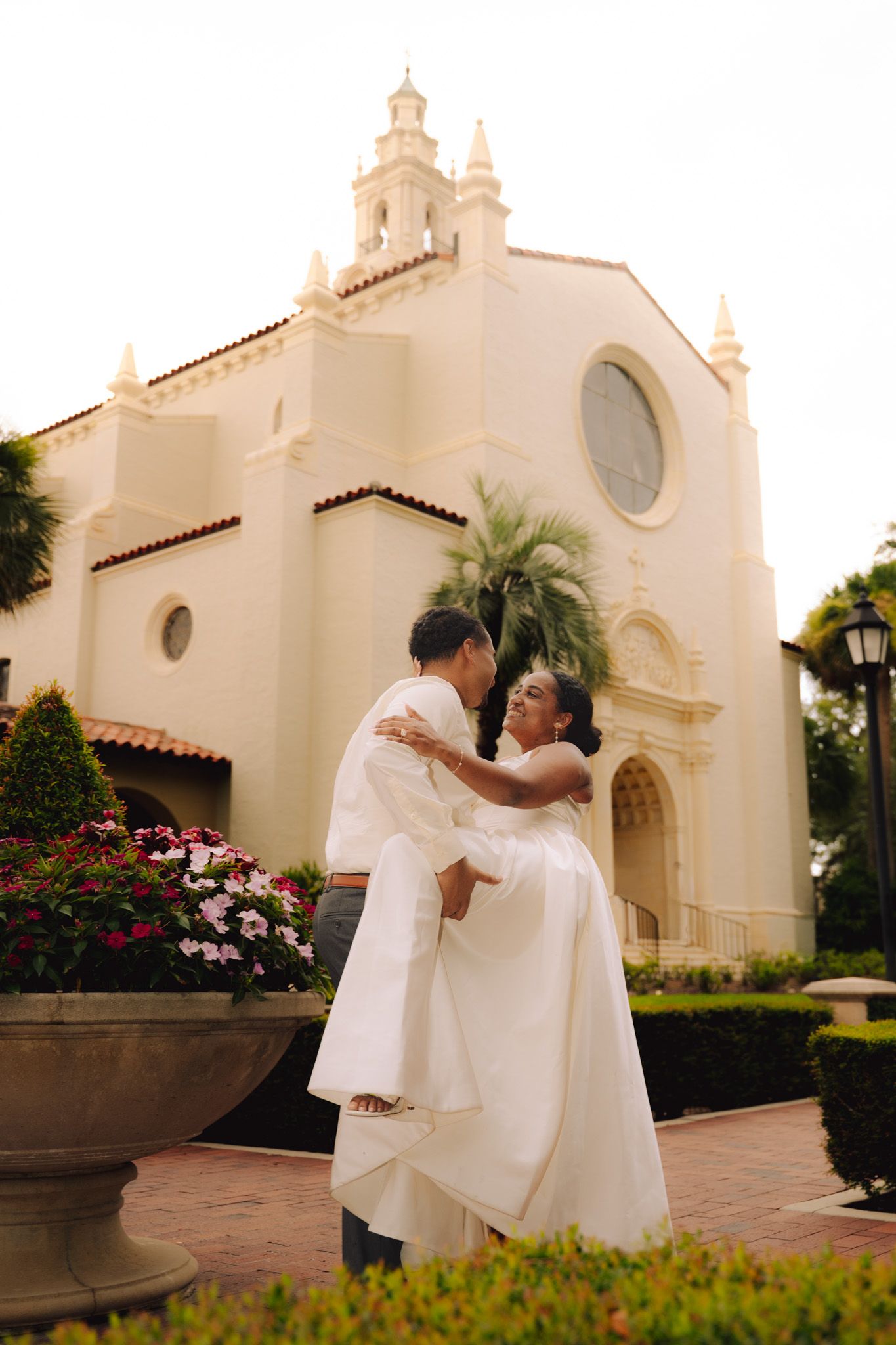Groom lifts bride in white dress outside elegant Spanish-style church surrounded by flowers.