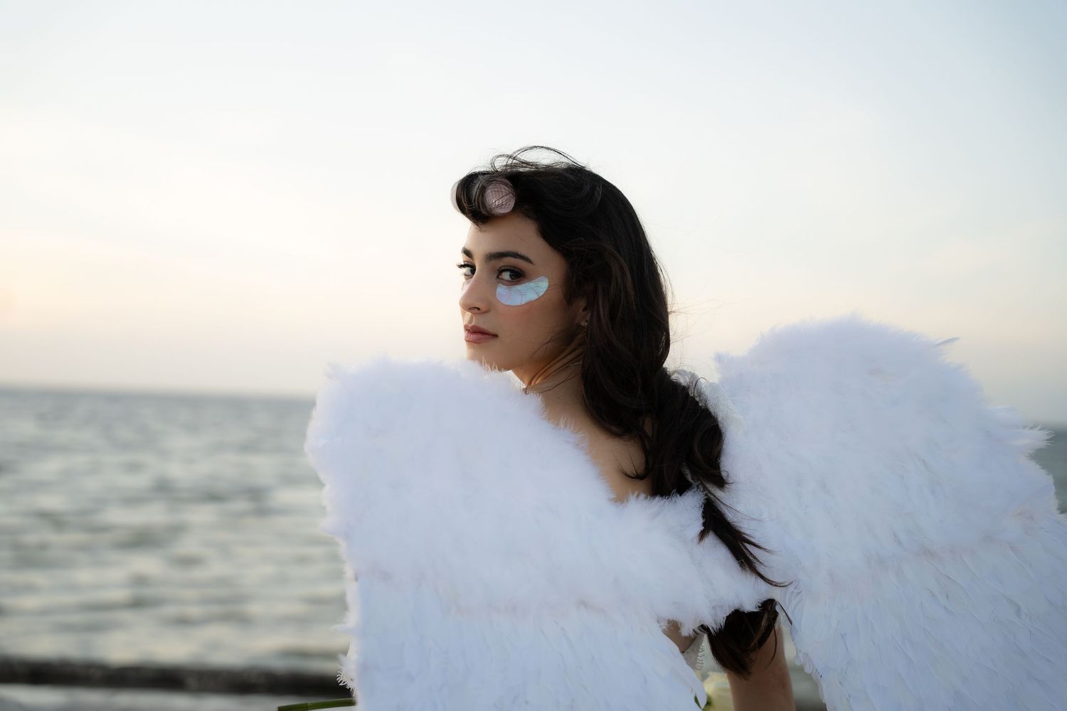 Young woman with angel wings and blue eye makeup gazing over her shoulder near the ocean.