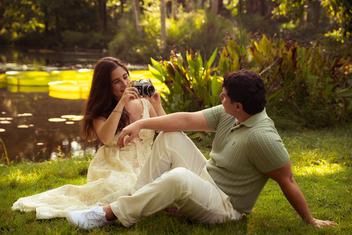 Couple sitting on grass by pond, man photographing woman in yellow dress with film camera.