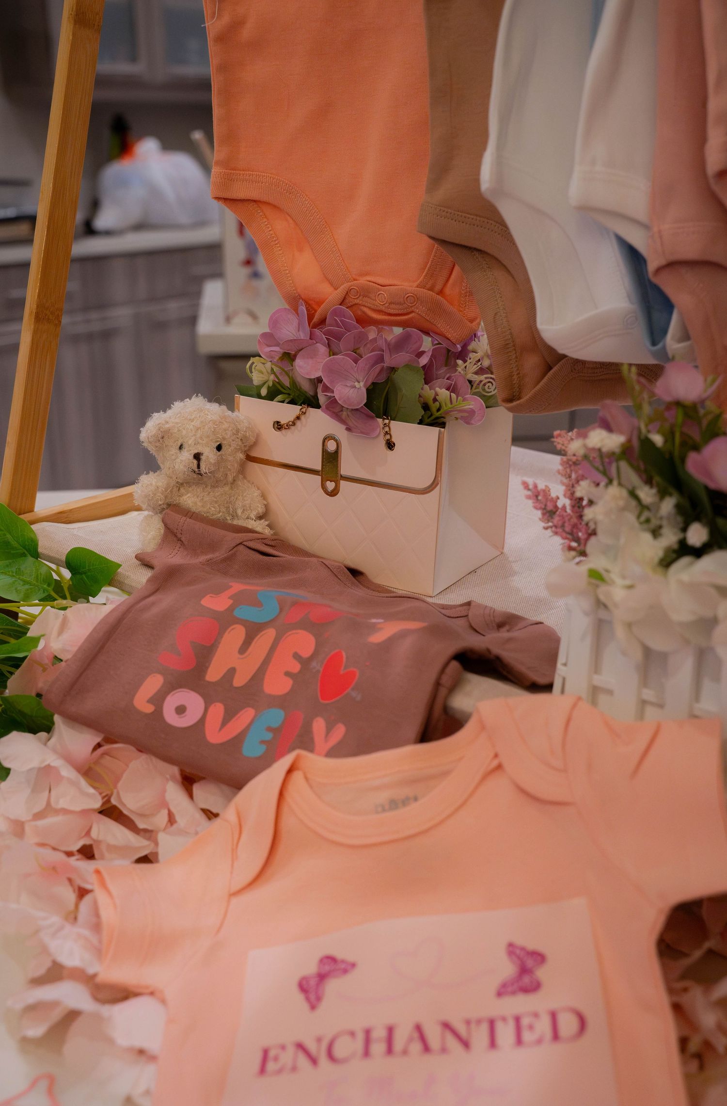 Pink and mauve baby onesies reading She Lovely and Enchanted displayed with teddy bear and flowers.