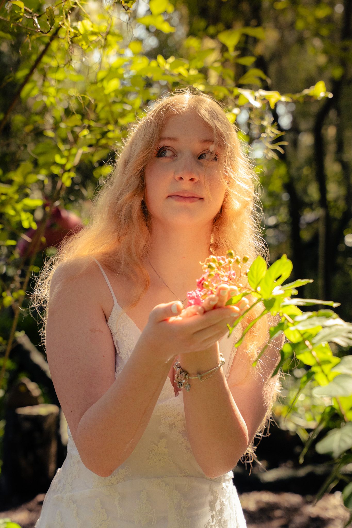Blonde woman in white lace dress holding pink flowers in a sunlit green forest.