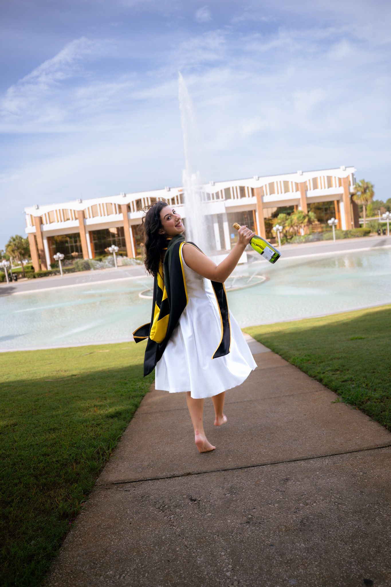 Joyful graduate in white dress and black gold regalia dances barefoot by university fountain.