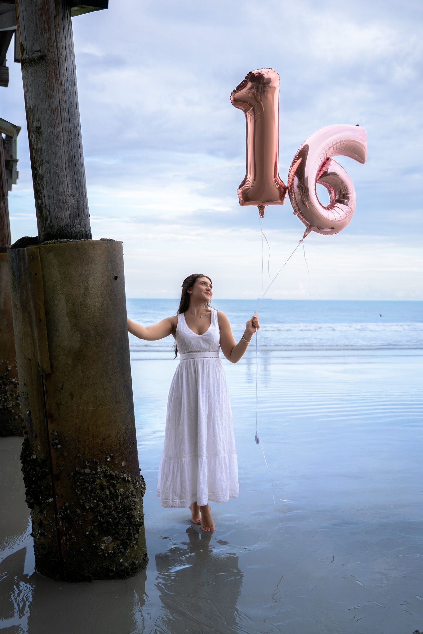Young woman in white dress holds rose gold number 16 balloons on a beach at low tide.