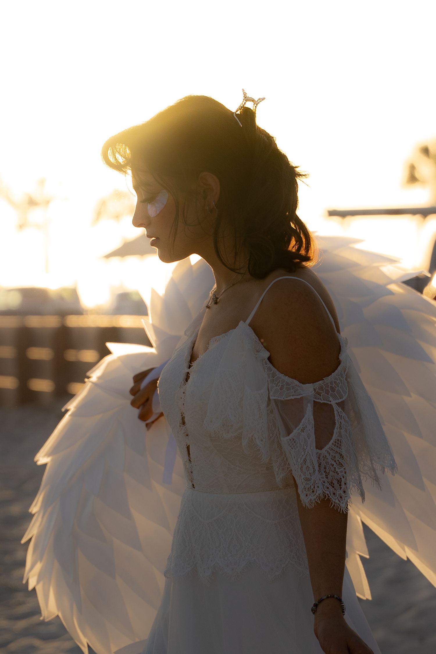 Woman in white lace dress with angel wings backlit by golden sunset light on a beach.