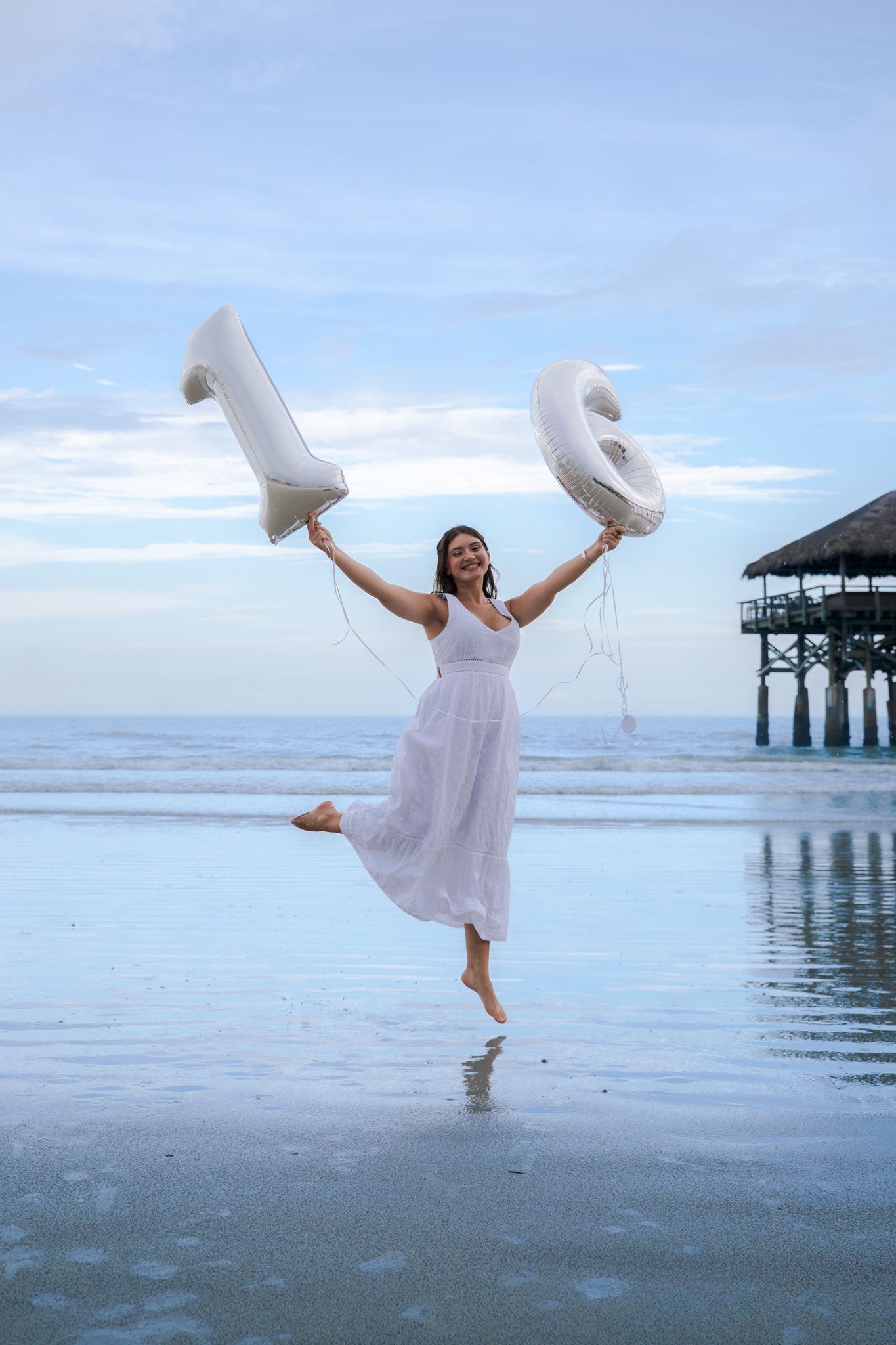 A joyful young woman in a white dress holds up number 16 balloons on a beach at dusk.