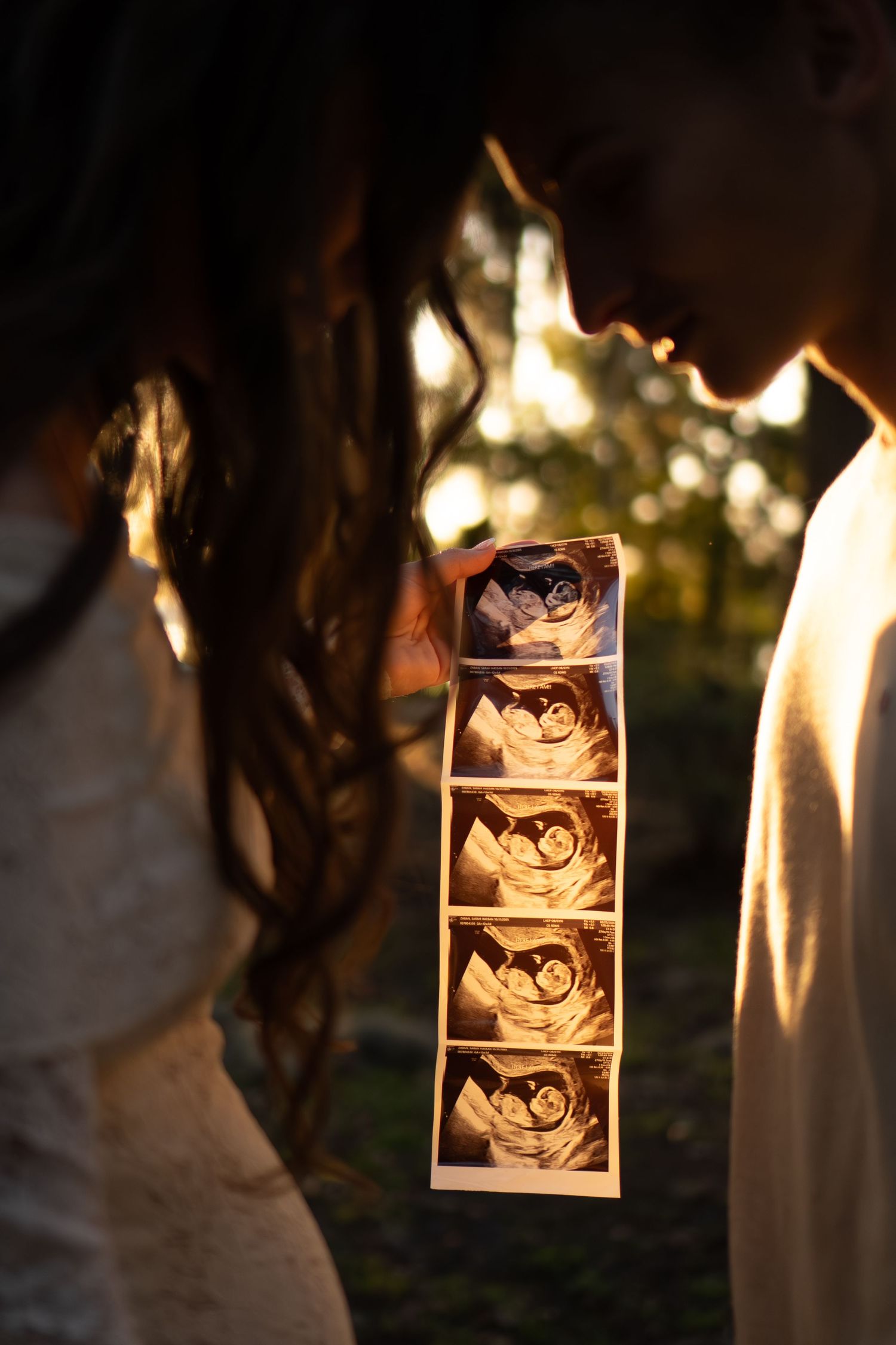 Couple holding ultrasound photos together in golden sunset light outdoors, sharing an intimate moment.