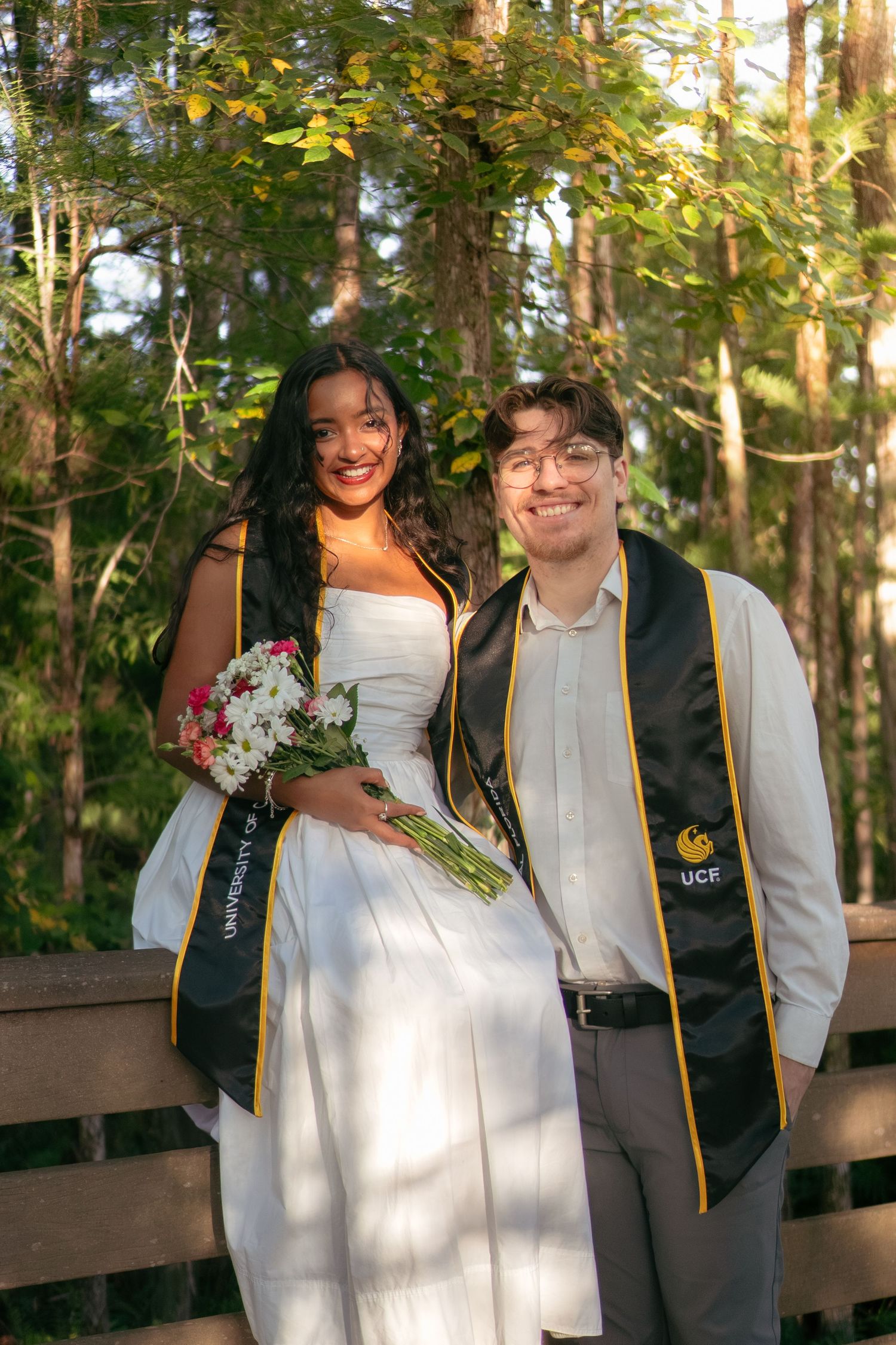Two UCF graduates in black and gold stoles pose together on a wooden bridge outdoors.