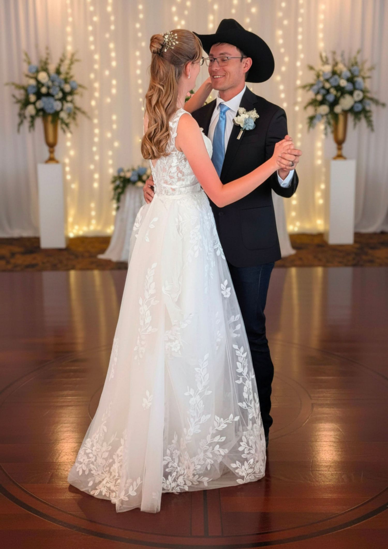 Bride and groom share first dance in elegant ballroom, bride in lace gown, groom in dark suit and cowboy hat.