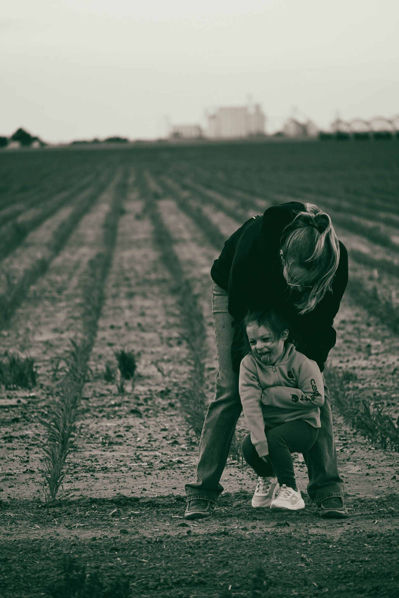 Black and white photo of mother bending over toddler in plowed farm field.
