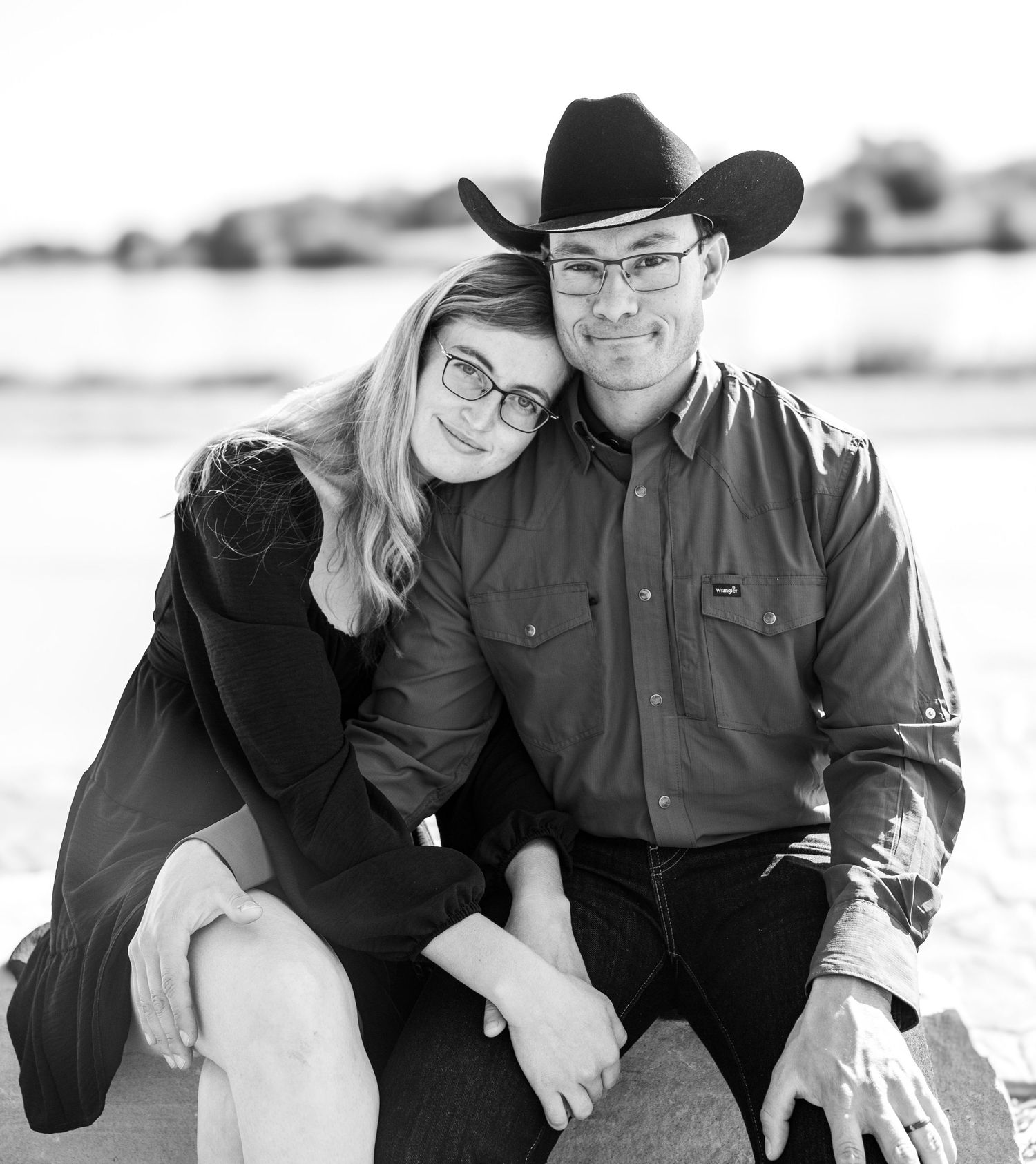 Black and white portrait of couple sitting together outdoors, man wearing cowboy hat by water.